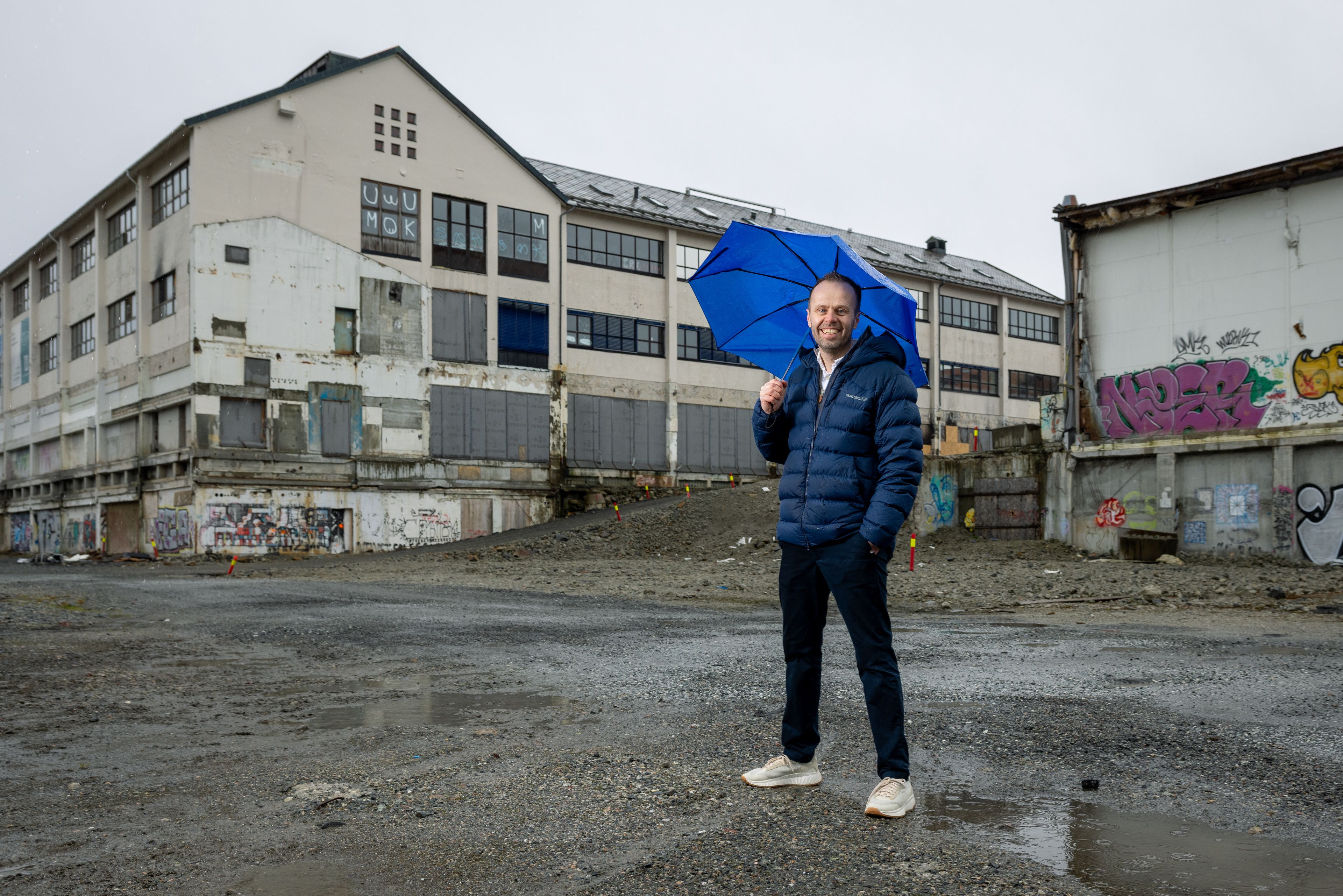 Prosjektleder Rønhovde står med paraply foran Tapperiet på byggetomten.