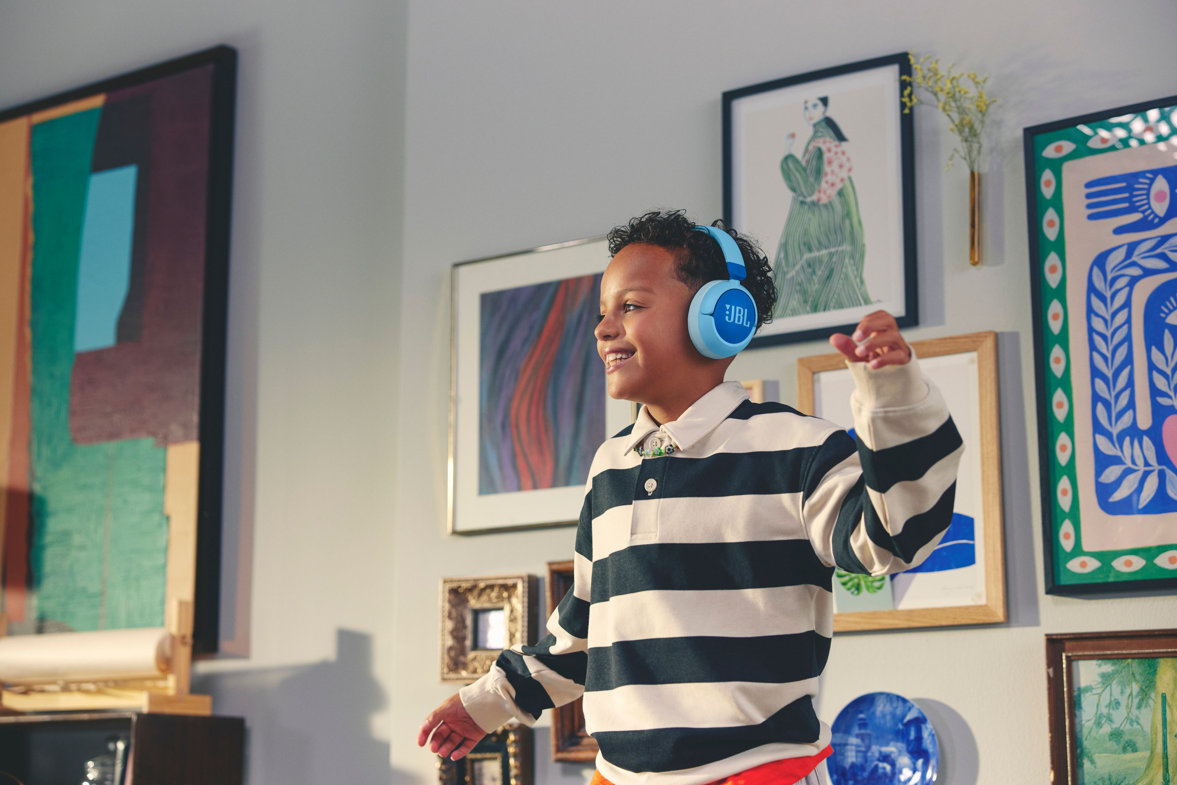 Et barn smiler bredt og danser litt med JBL hodetelefoner på hodet. Det er masse kunstverk på veggene bak han.