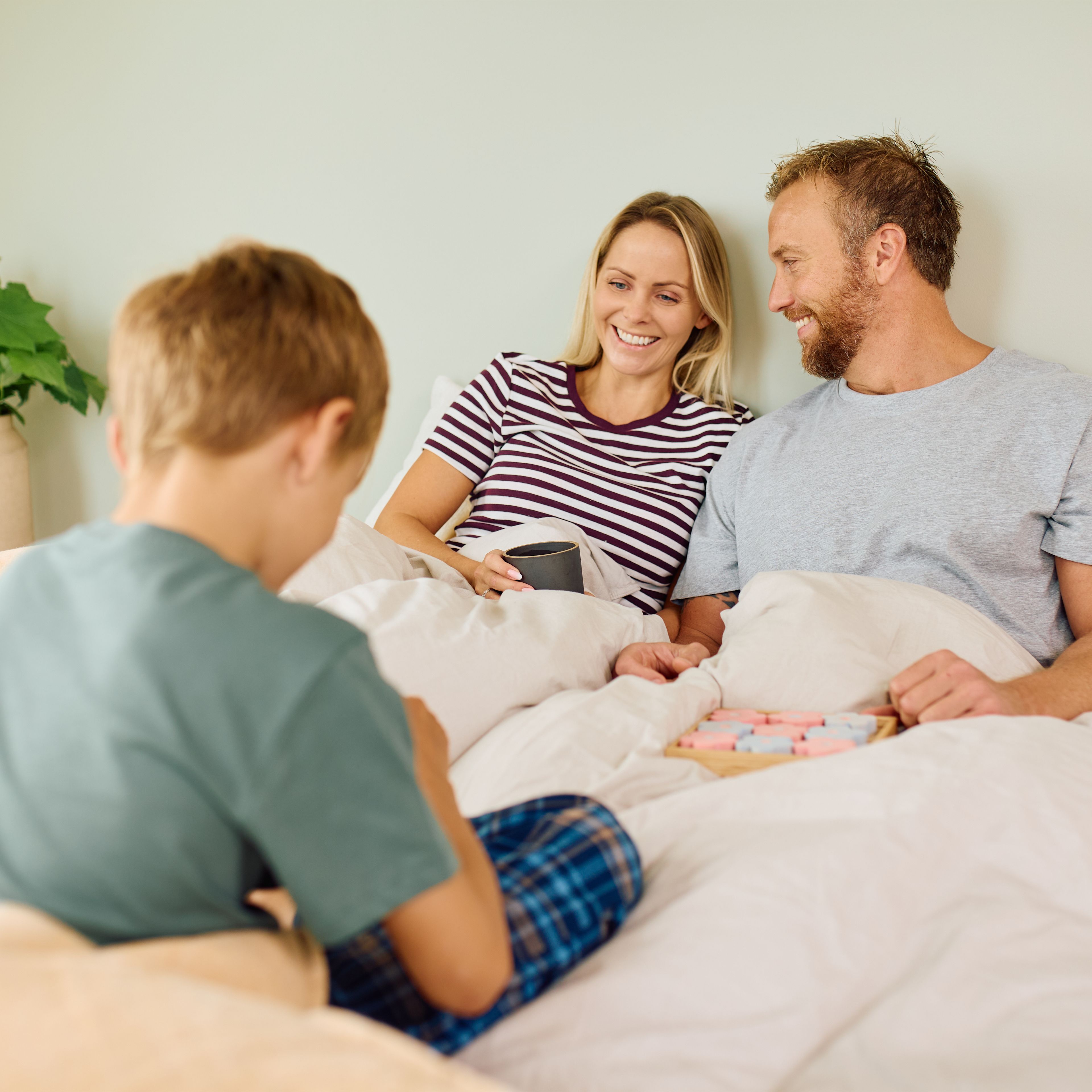 En familie sitter i sengen. En mann og kvinner smiler til hverandre. En gutt sitter også i sengen.