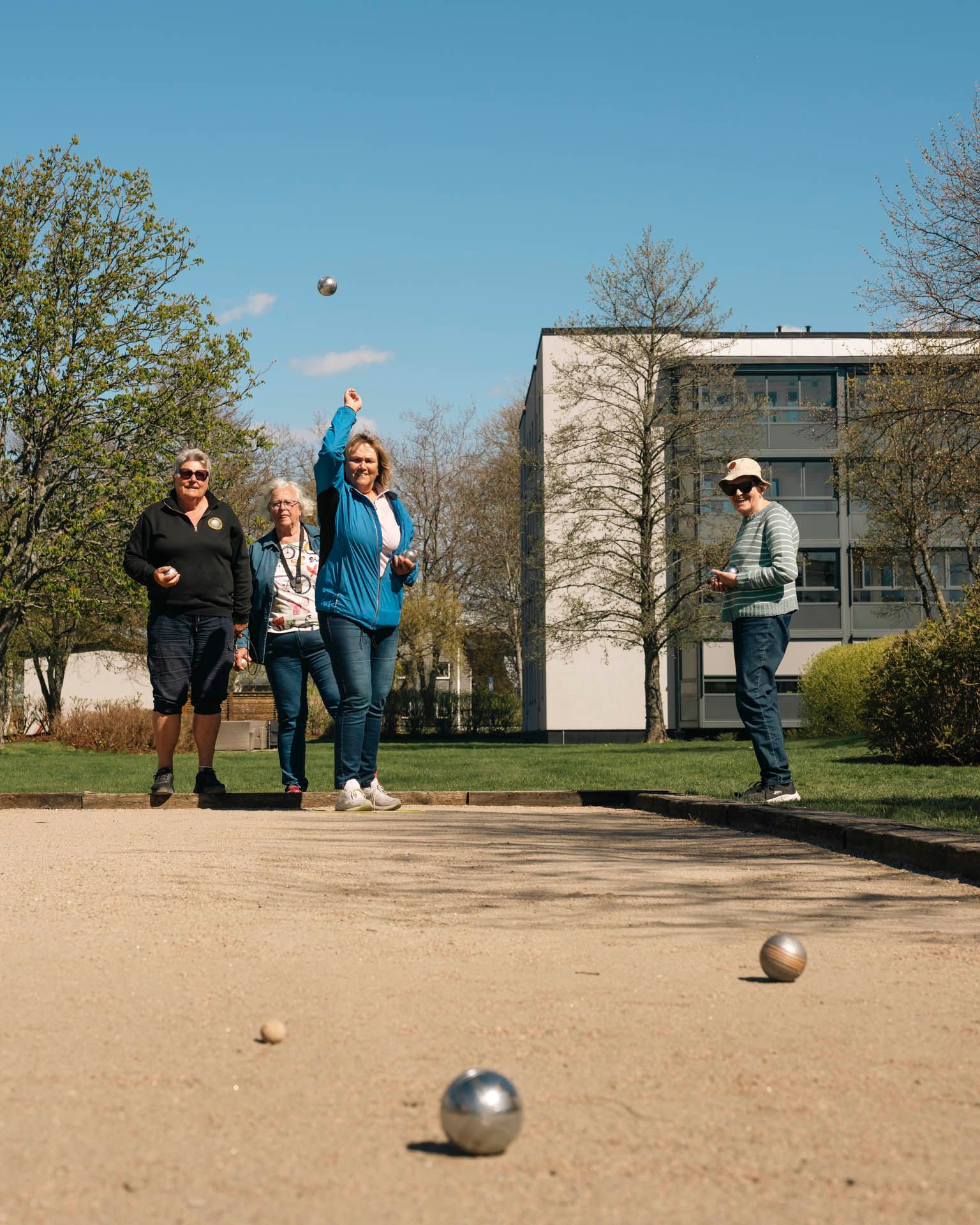 Flere personer spiller petanque på en grusbane mellom boligblokker en solrik dag.