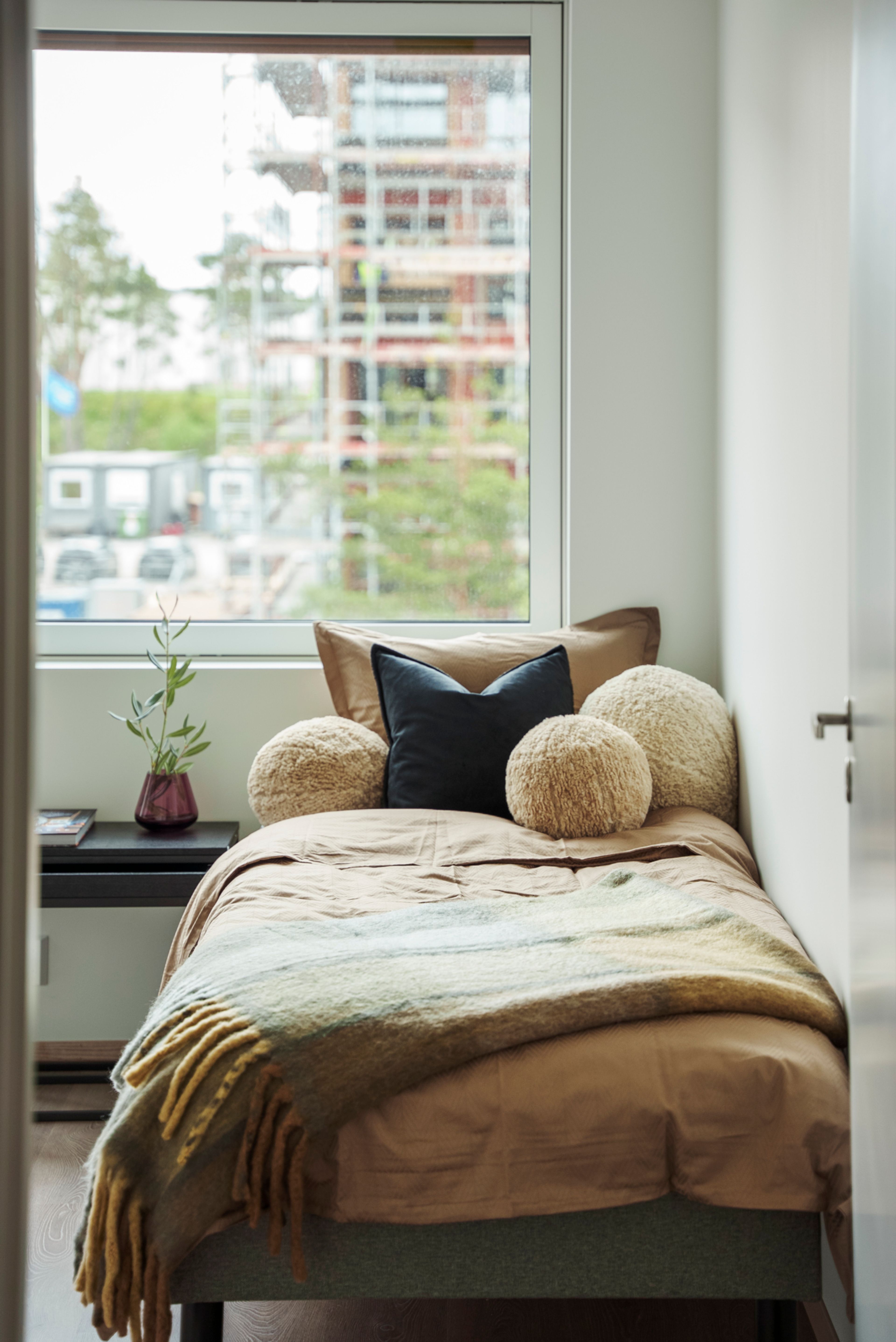 Et soverom med enkeltseng og et stort vindu, dekorert med beige sengetøy og sort nattbord.