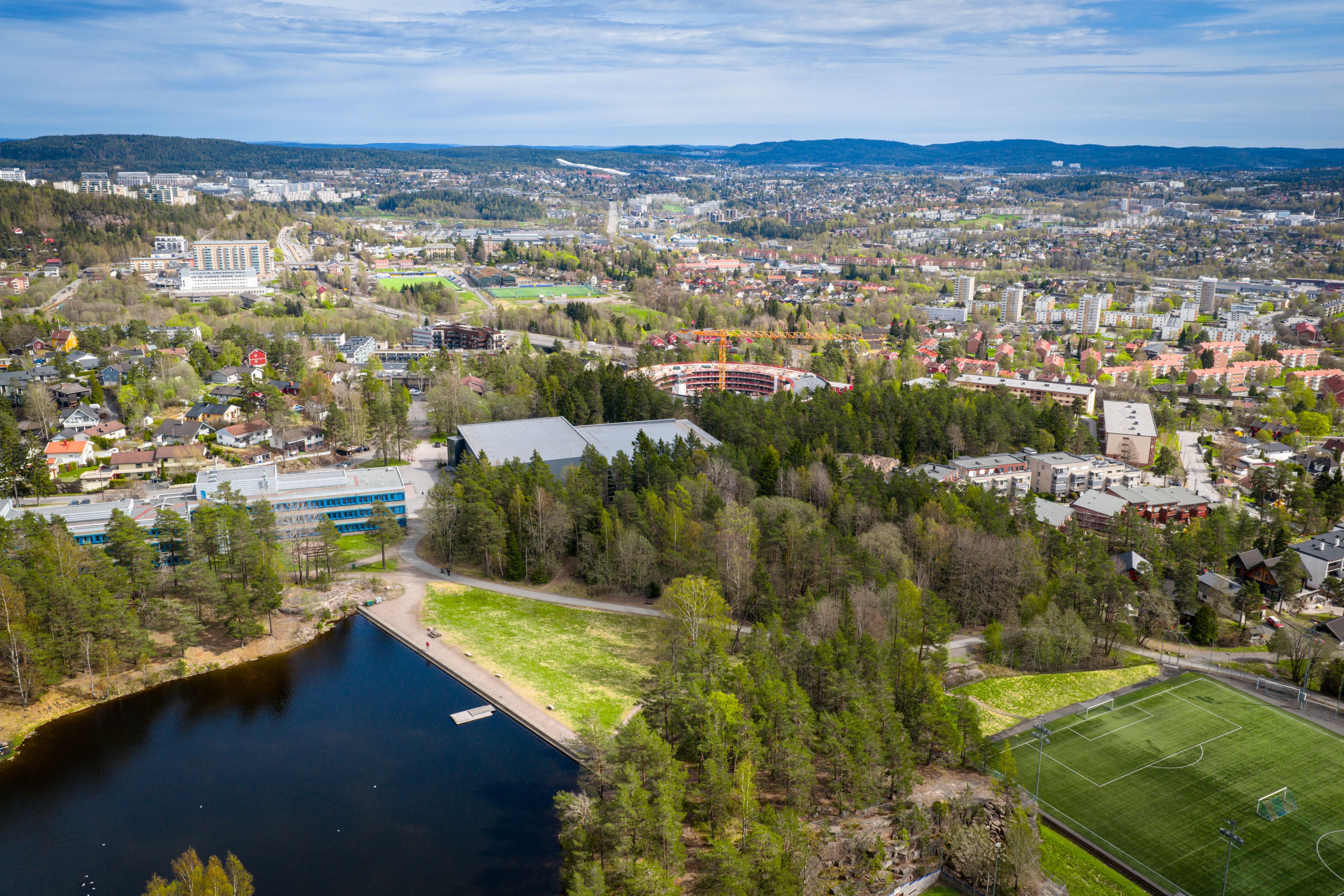 Dronefoto over Rødtvet i Oslo. Midt i bildet synes det sirkelformede Oen-prosjektet under oppføring, med en gul byggekran. Til venstre ligger Vesletjern med badestrand og en skole med blå fasade. Til høyre en fotballbane med kunstgress. Rundt ligger villabebyggelse og boligblokker, med Oslos åser i horisonten.
