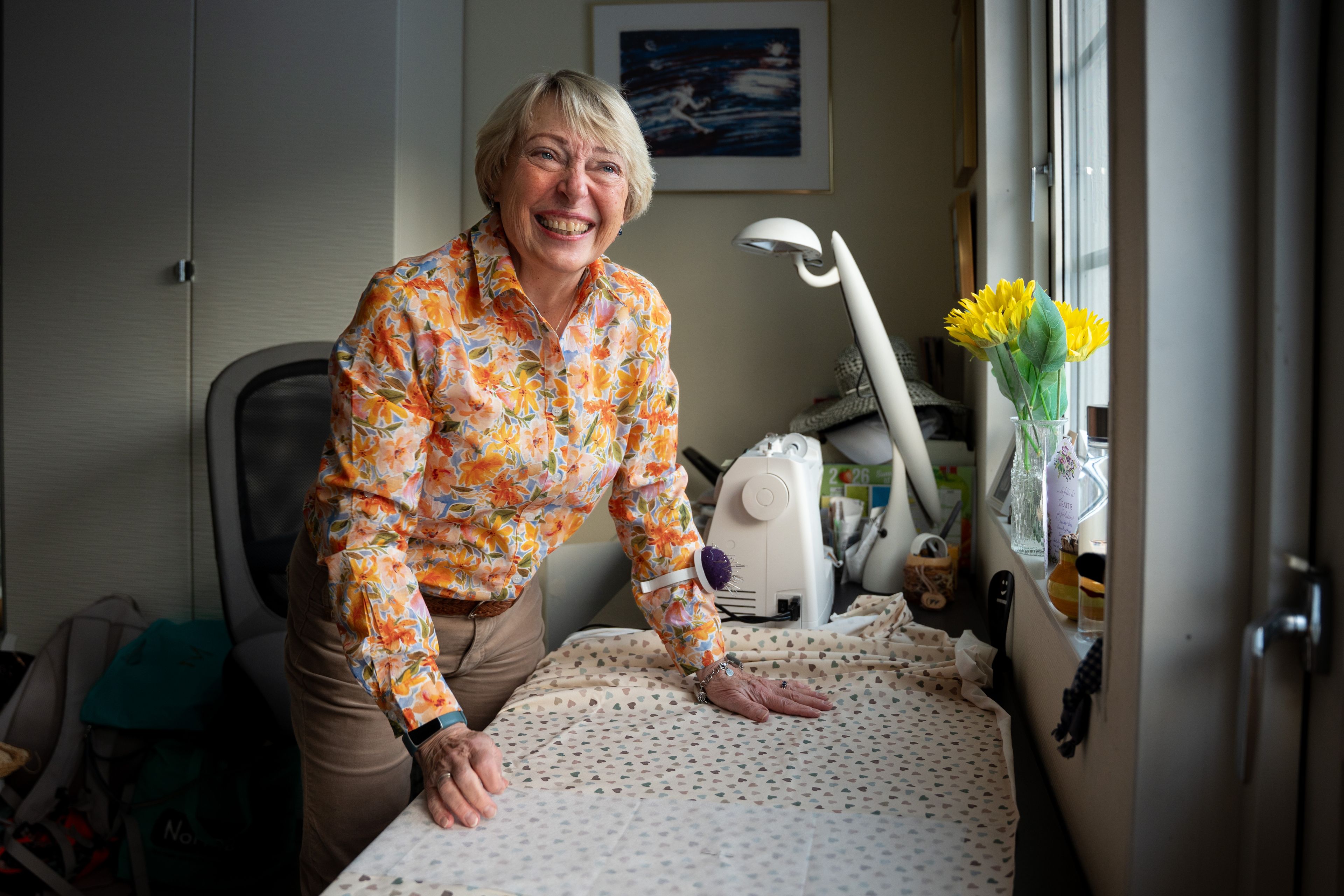 Smilende kvinne i blomstrete skjorte står ved sybordet og arbeider med stoff foran symaskinen i et lyst hobbyrom.