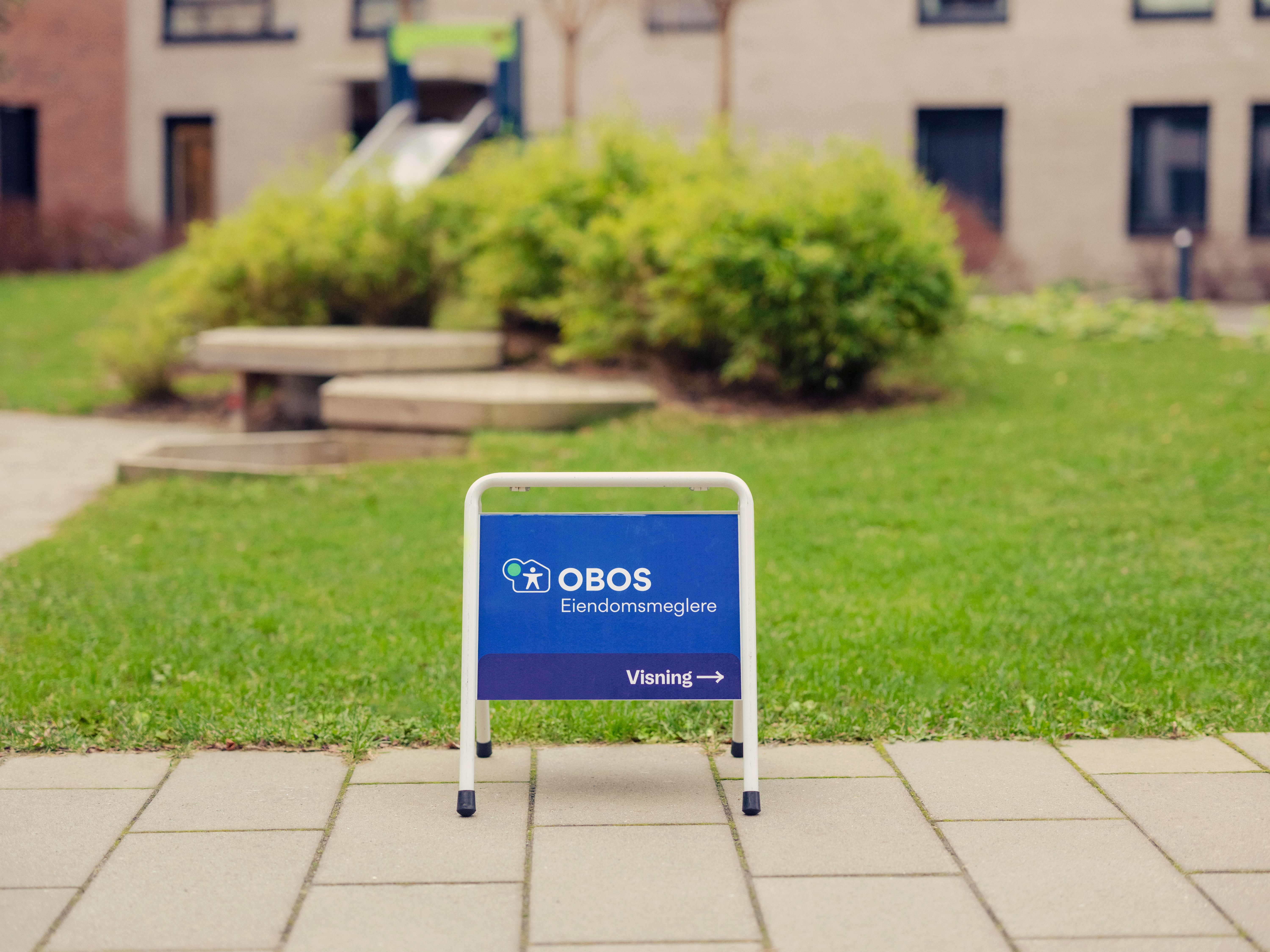 Et OBOS Eiendomsmeglere-skilt står på bakken foran en grønn plen.