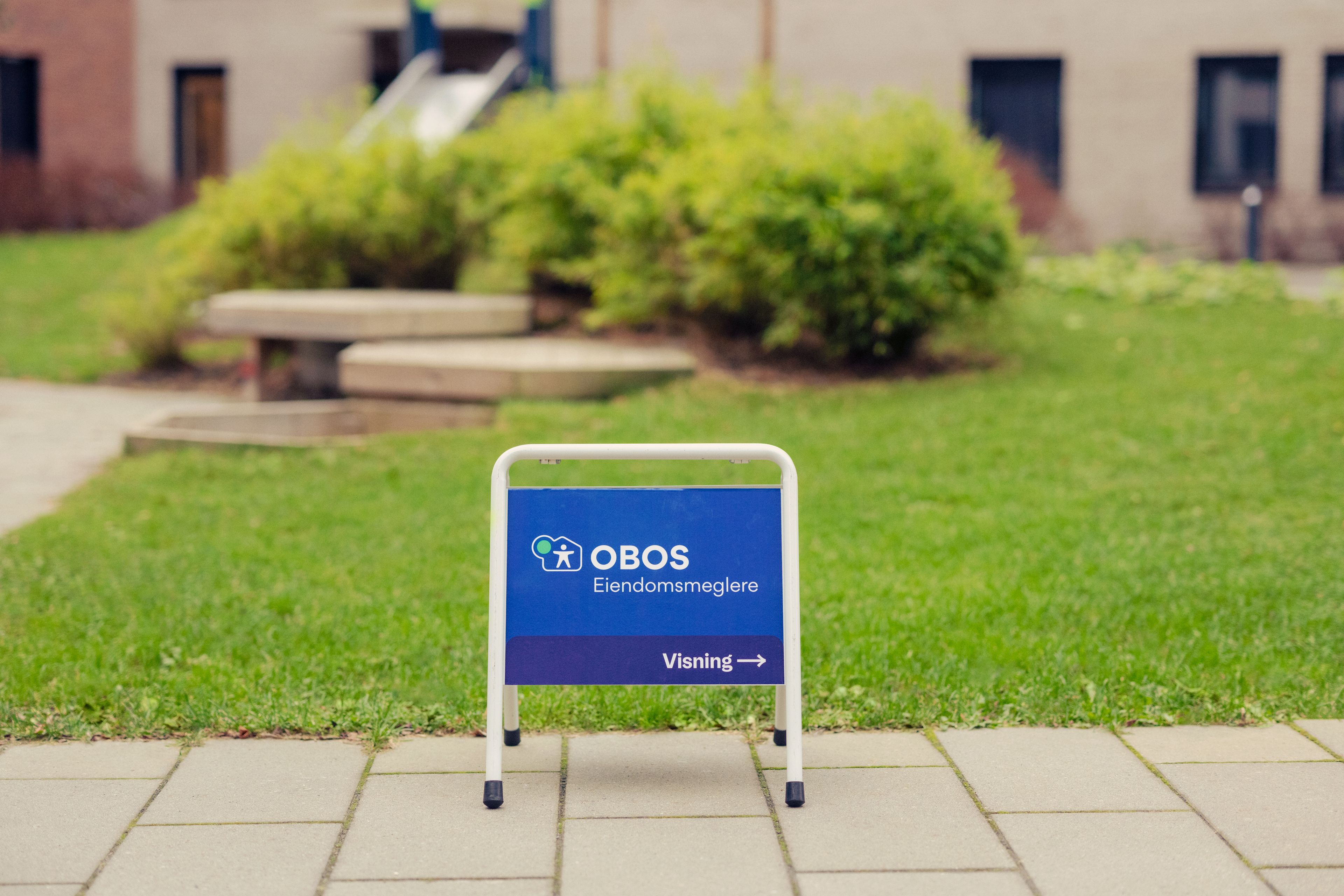 Et OBOS Eiendomsmeglere-skilt står på bakken foran en grønn plen.