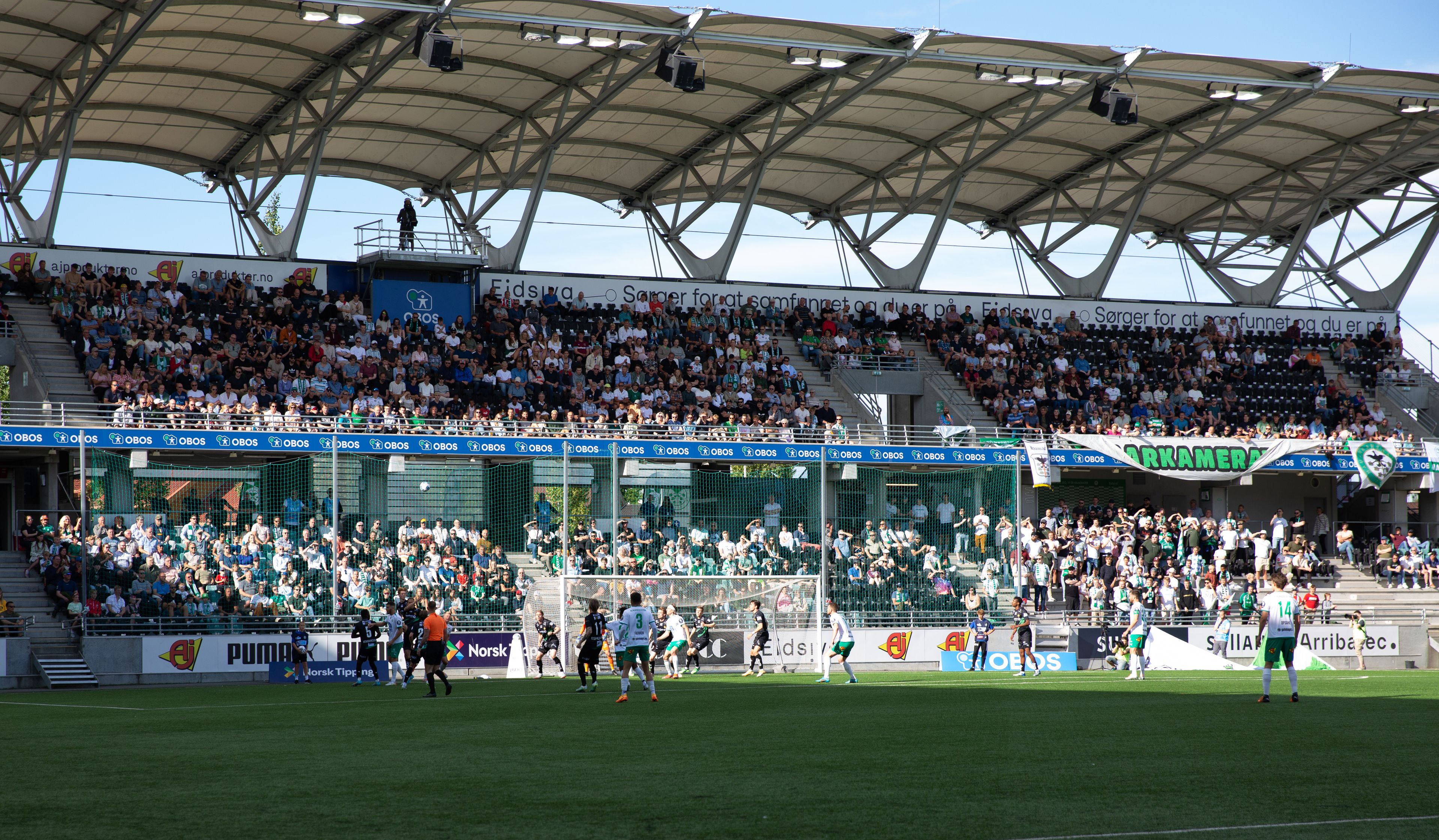 Bilde av HamKam supportere som står på tribunen rundt fotballbanen og heier på fotballspillerne på banen.