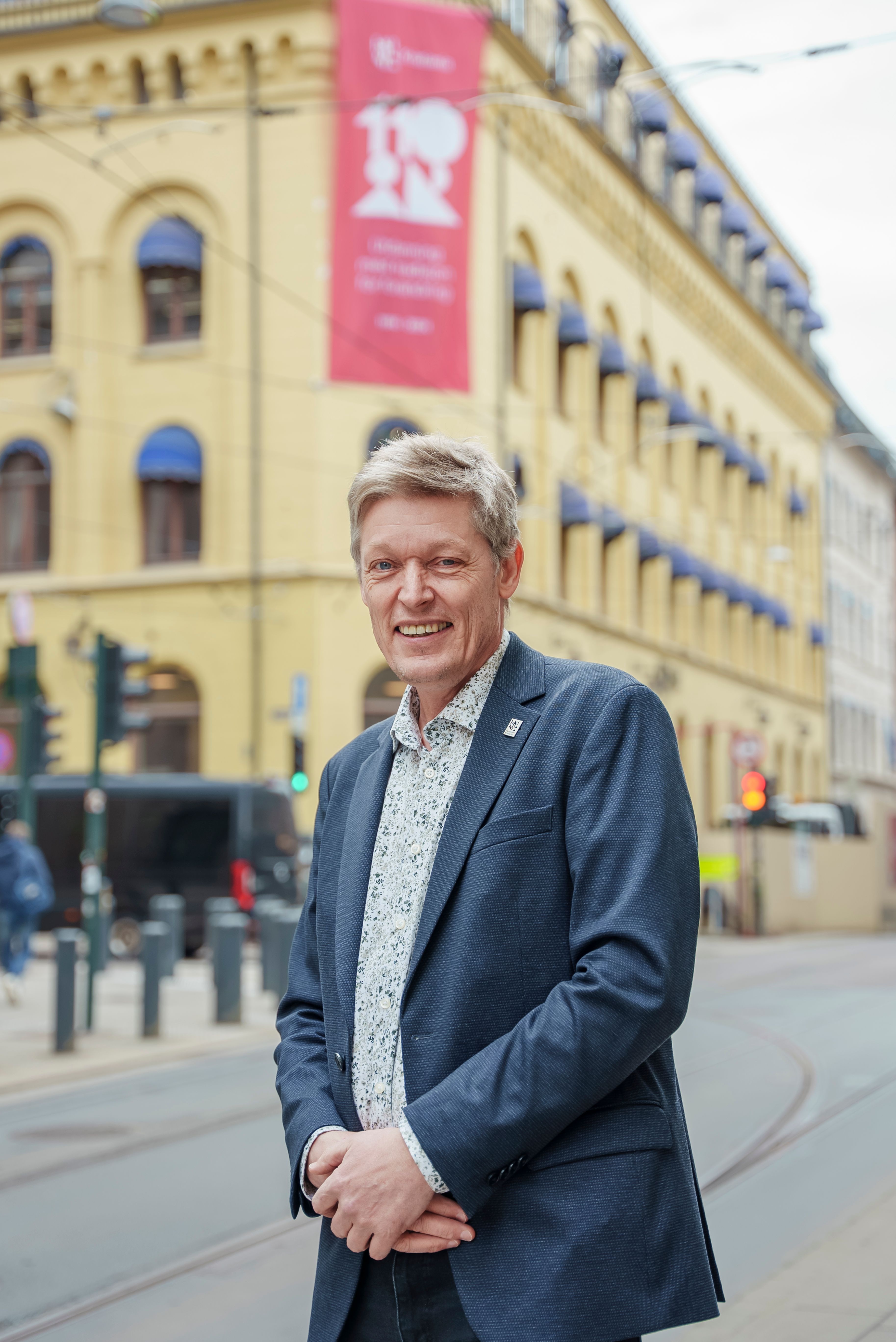 En mann står utenfor bygningen til Høyskolen Kristiania i Kirkegata. Han har på seg dressjakke. Han smiler bredt.