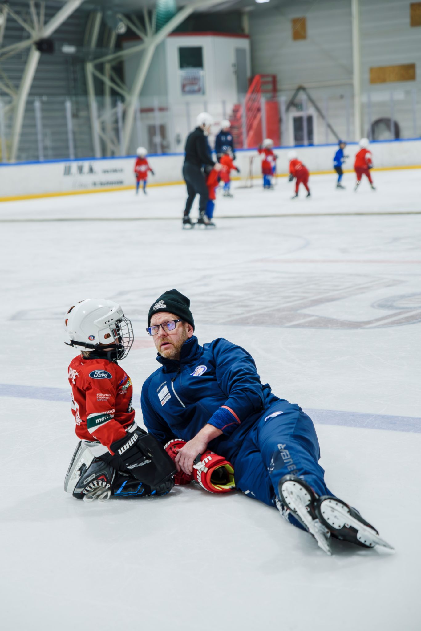 Foto av skøytetreneren som har lagt seg ned på isen for å snakke med et av barna på skøyteskolen.