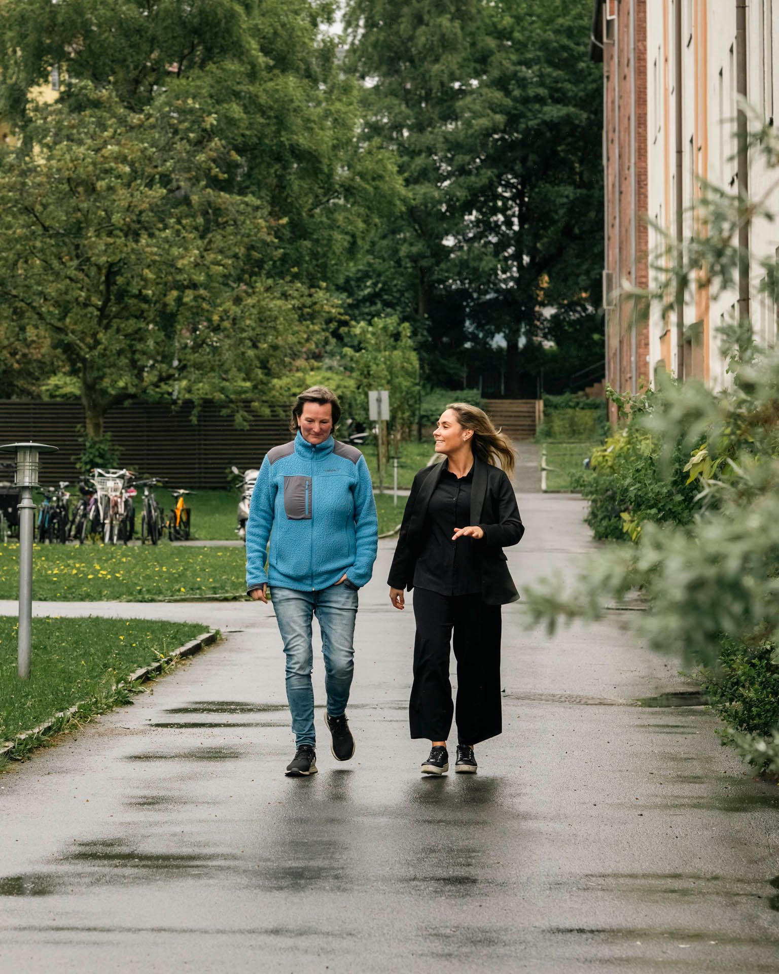 To personer går sammen på en våt gangvei ved Gjøvikgata borettslag, omgitt av grønt løvverk og boligblokker.