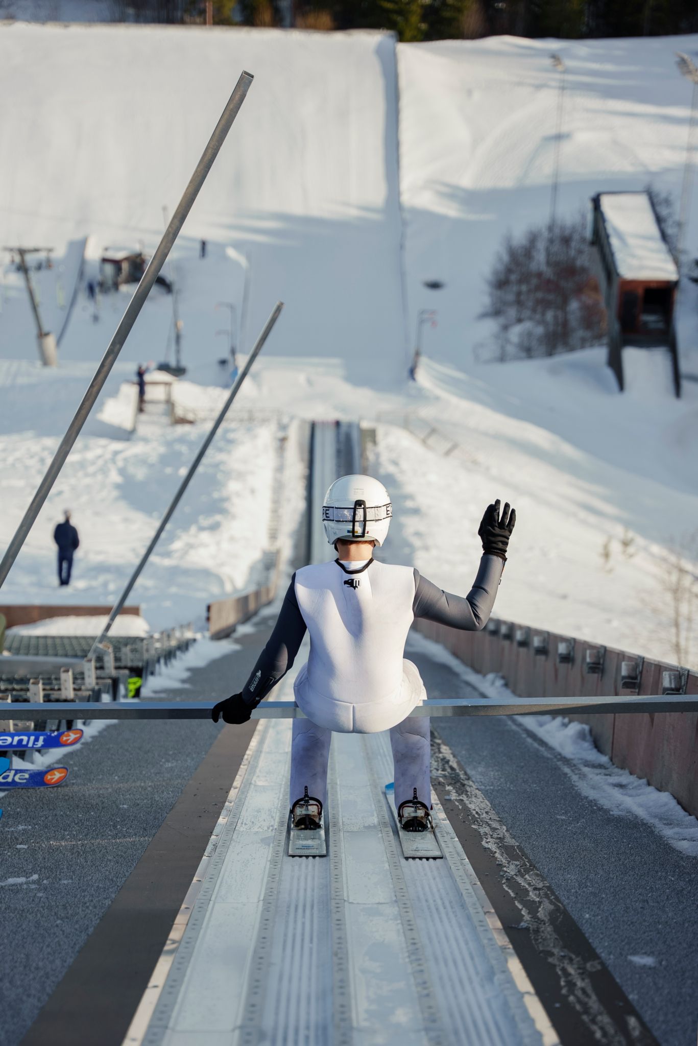 En ungdom sitter på bommen med utsikt utover skihoppet. Vedkommende er filmet bakfra og har høyre hånd hevet.