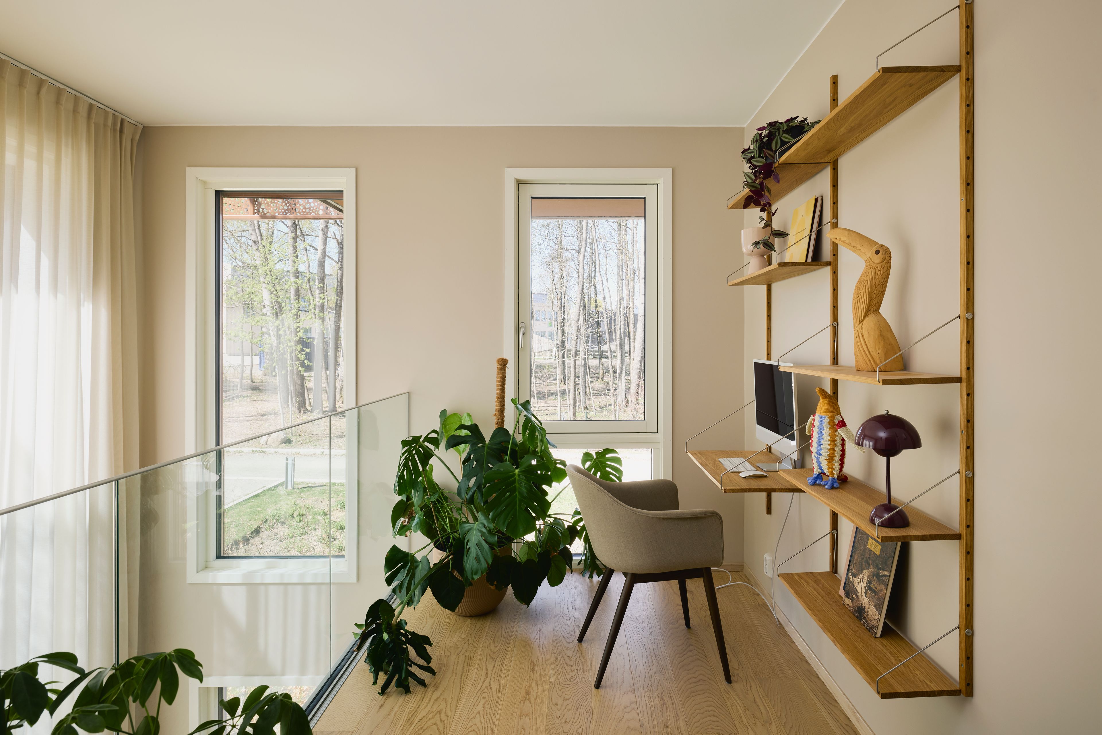 Kontorkrok i andre etasje med veggmontert trehylle, skrivepult, beige stol, store monstera-planter og to høye vinduer mot skogen.
