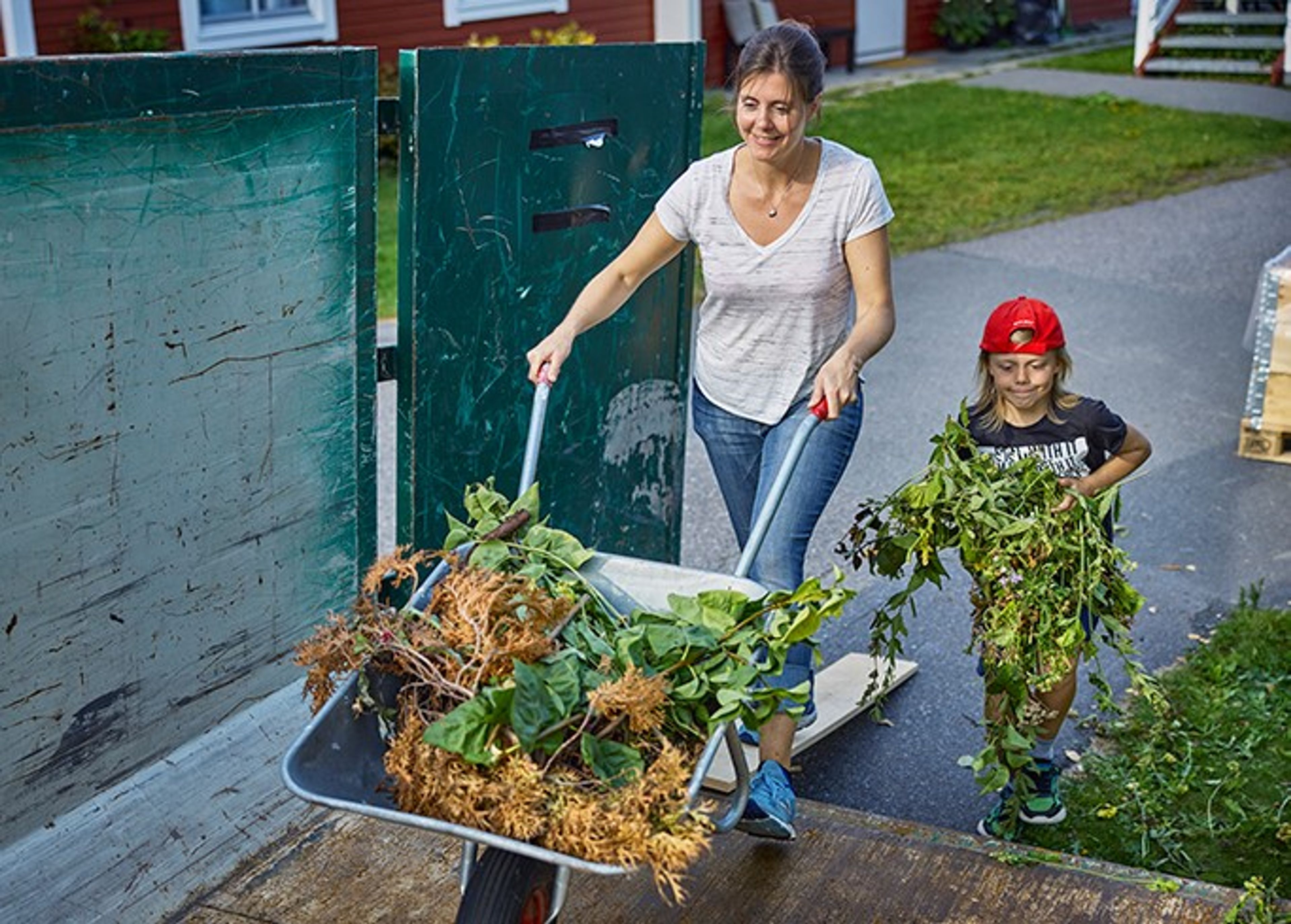 Ei dame triller en trillebår med hageavfall, og en liten gutt hjelper henne.