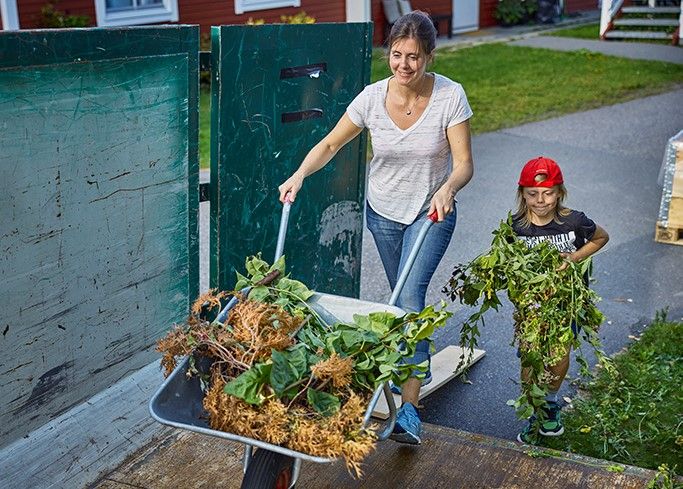 Ei dame triller en trillebår med hageavfall, og en liten gutt hjelper henne.