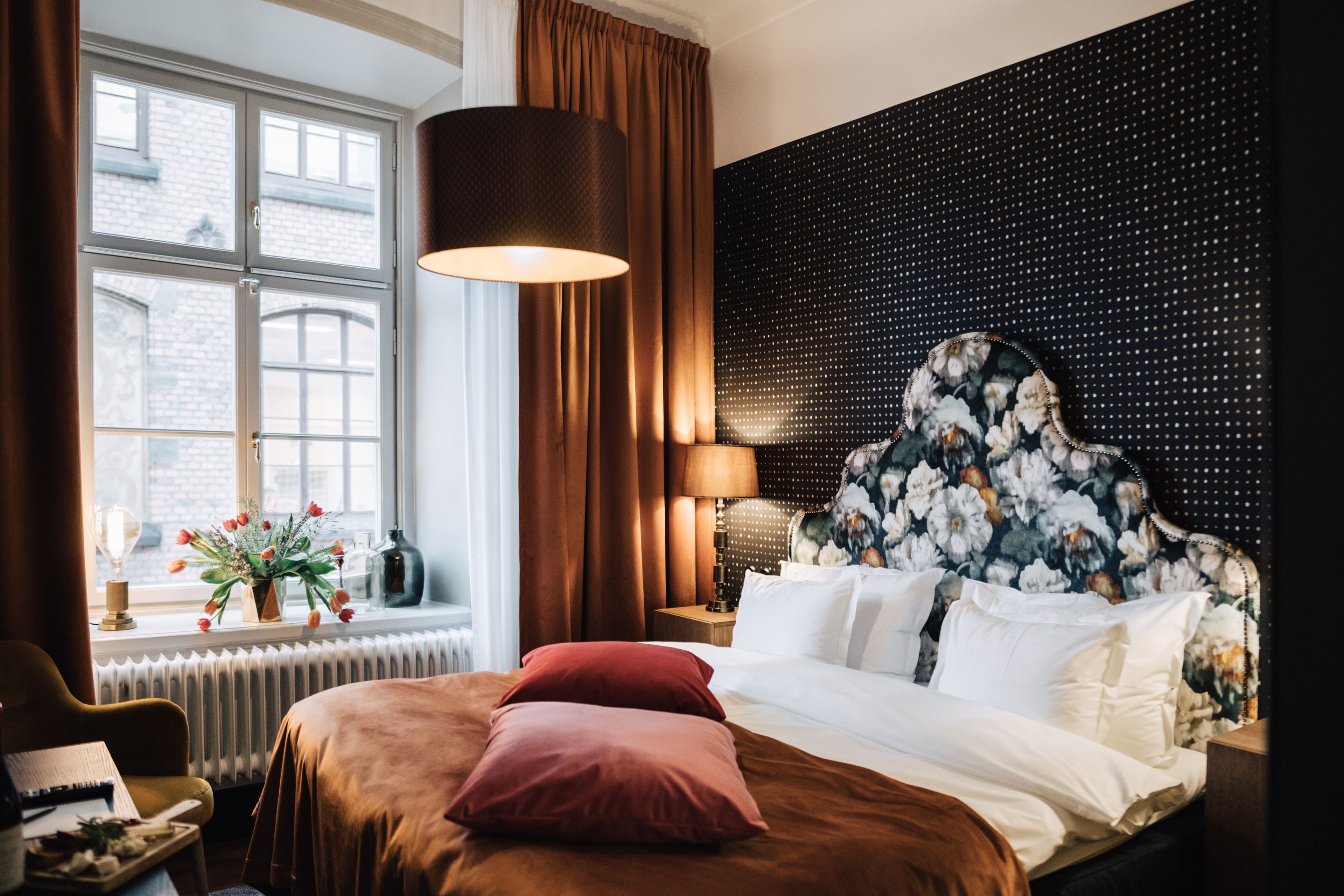 Bilde fra et hotellrom. Det er ganske innbydende utseende med blomster i vinduskarmen og en stor deilig seng.
