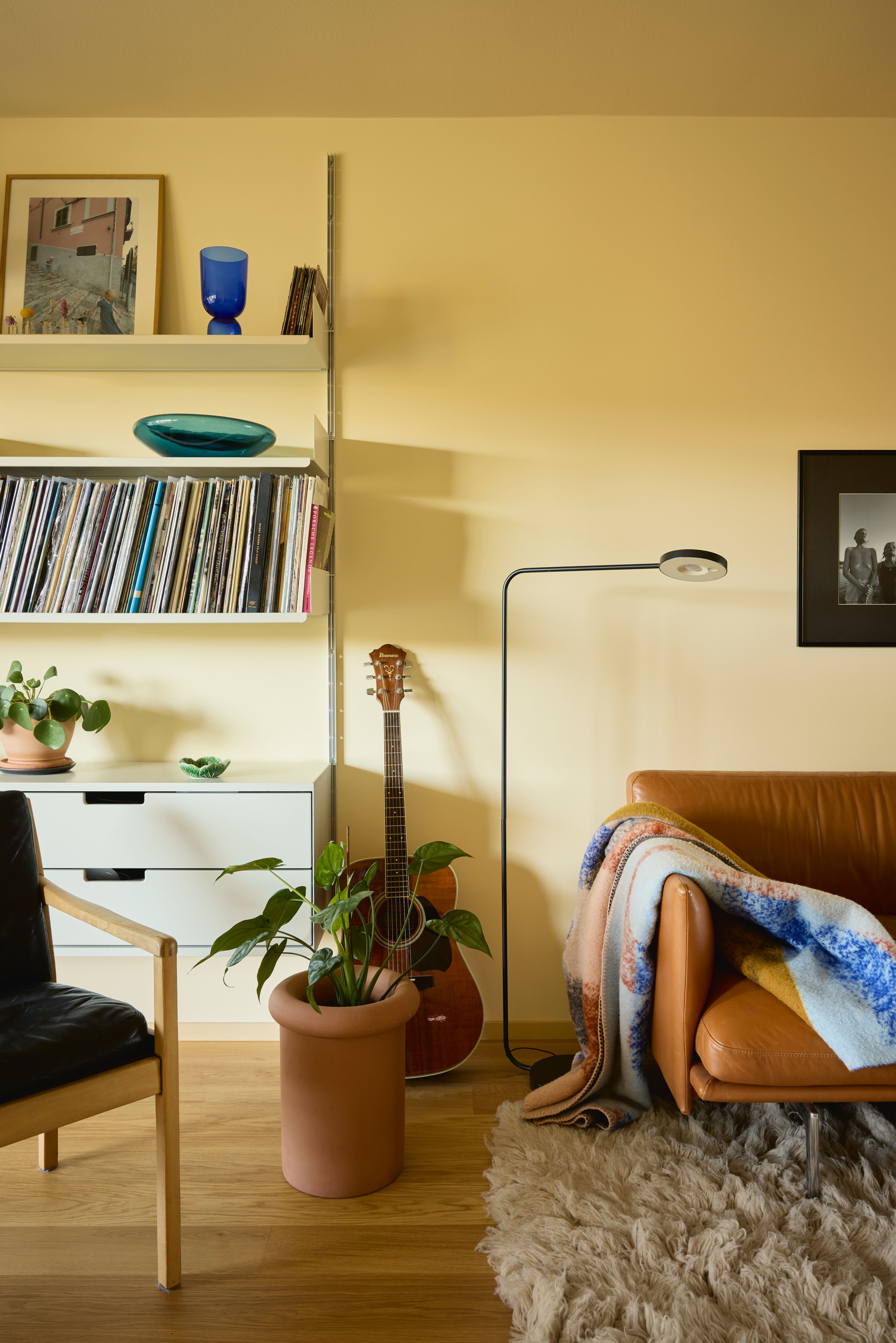 Nærbilde fra stua av sofa med et lunt teppe, grønn plante og en vegghengt hylle med bøker.