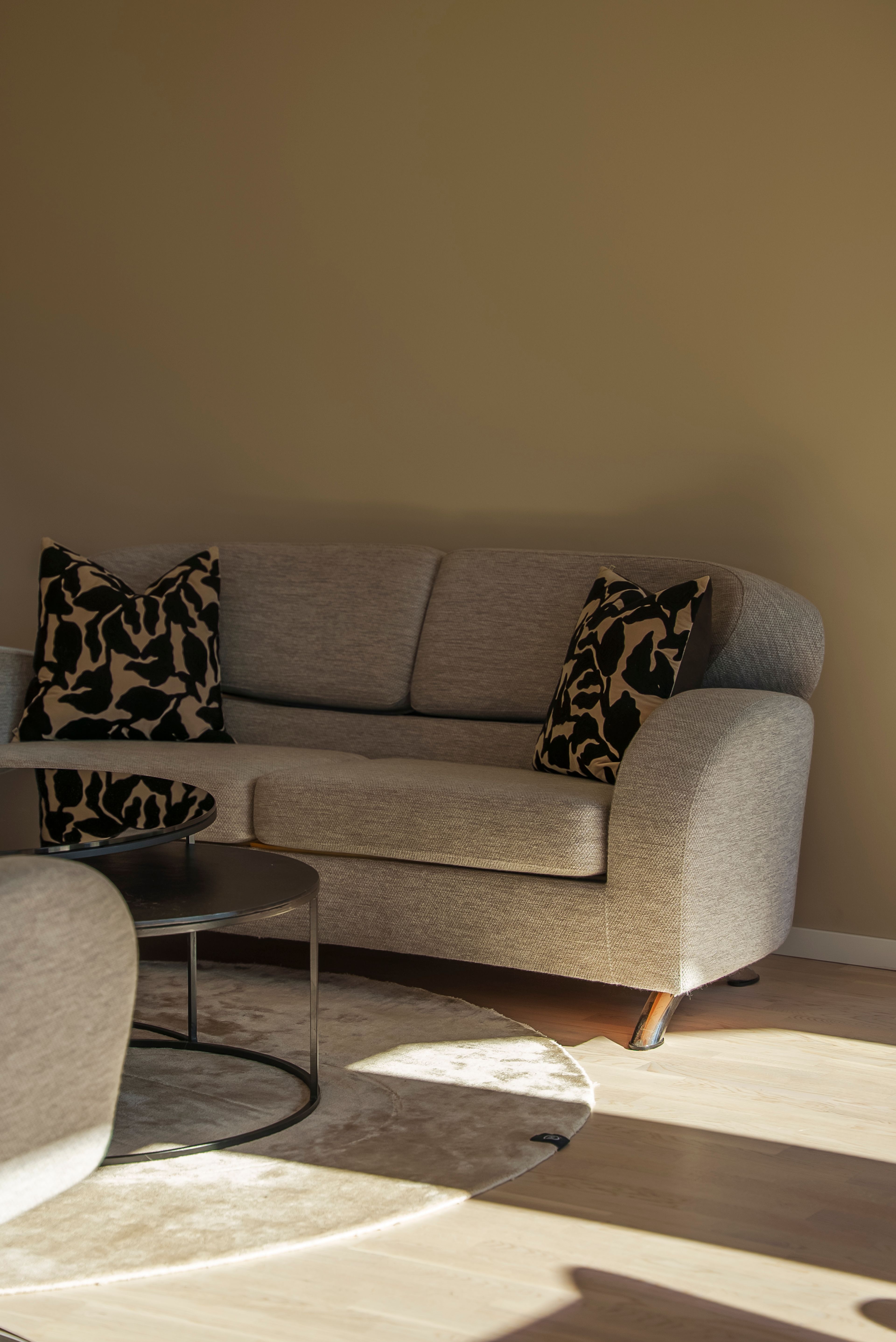 Nærbilde av en grå sofa med noen beige puter med svarte blader på, mot en beige vegg