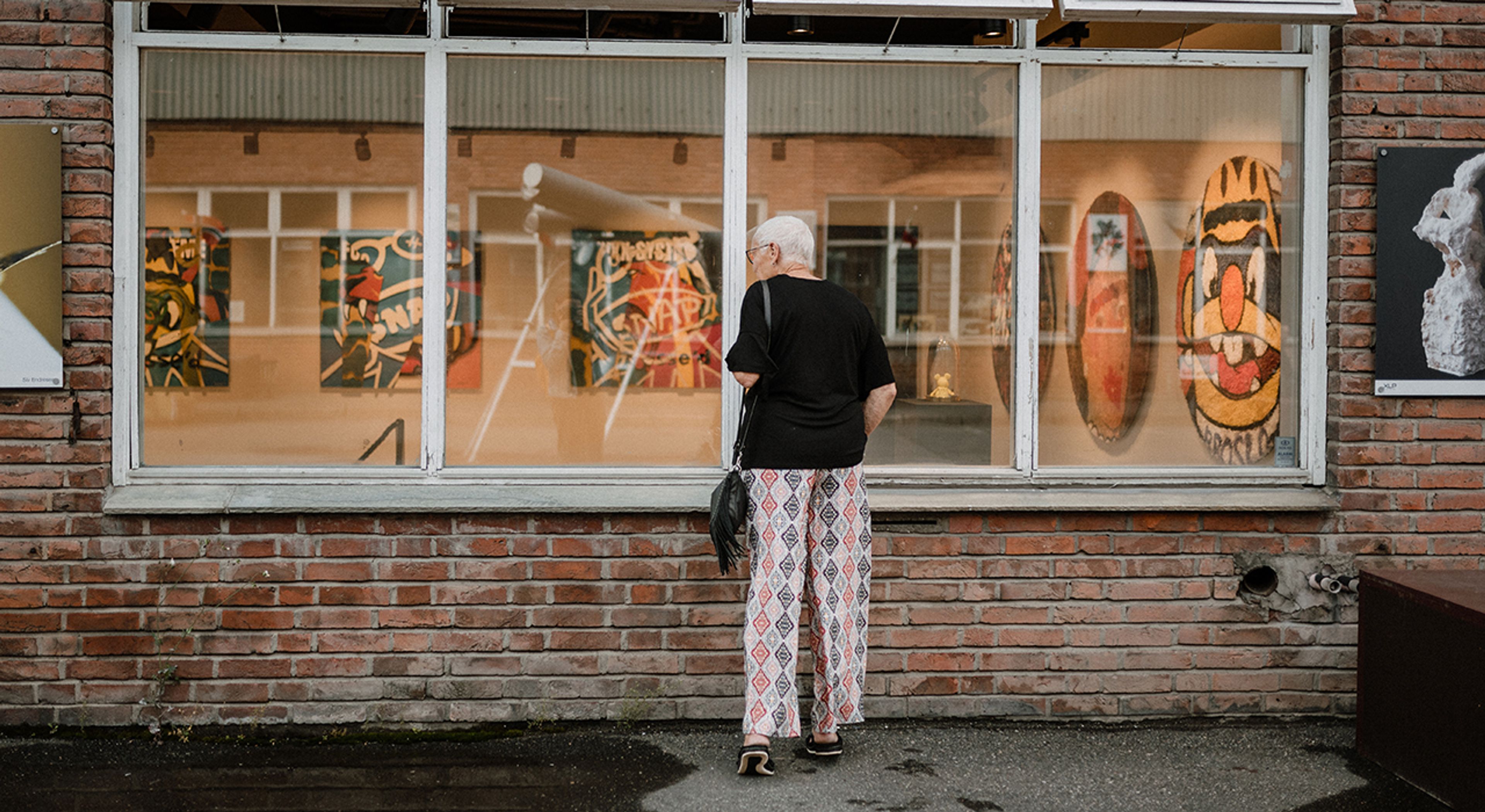 Eldre kvinne som titter inn i vinduet til et kunstgalleri på dagtid.