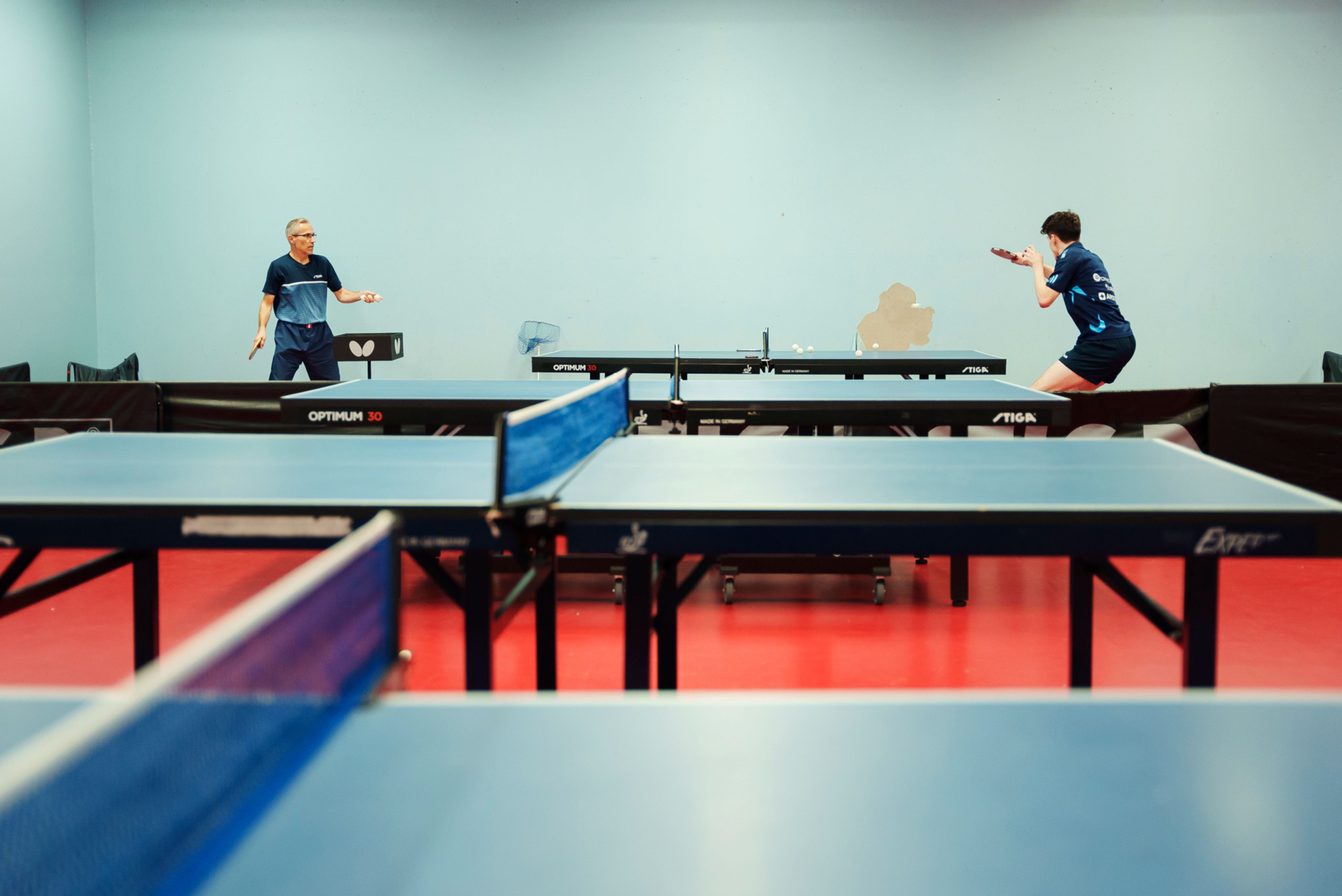 Michael og en trener står på hver sin side av et bordtennisbord mot en blå vegg. I forgrunnen to andre bordtennisbord.