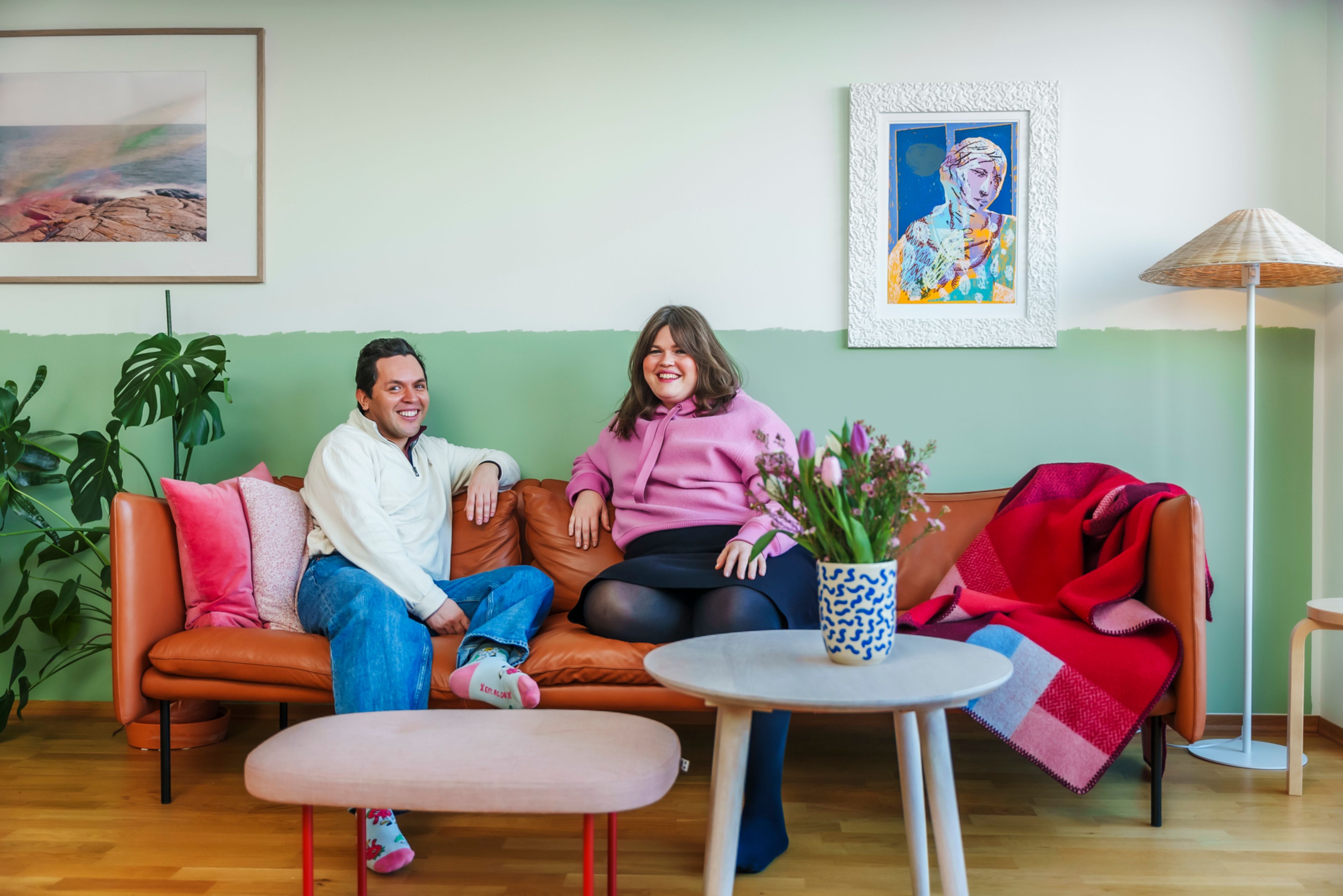 Et vennepar som sitter i en brun skinnsofa i en fargerik leilighet med grønne vegger, grønne planter og rød sittepuff.