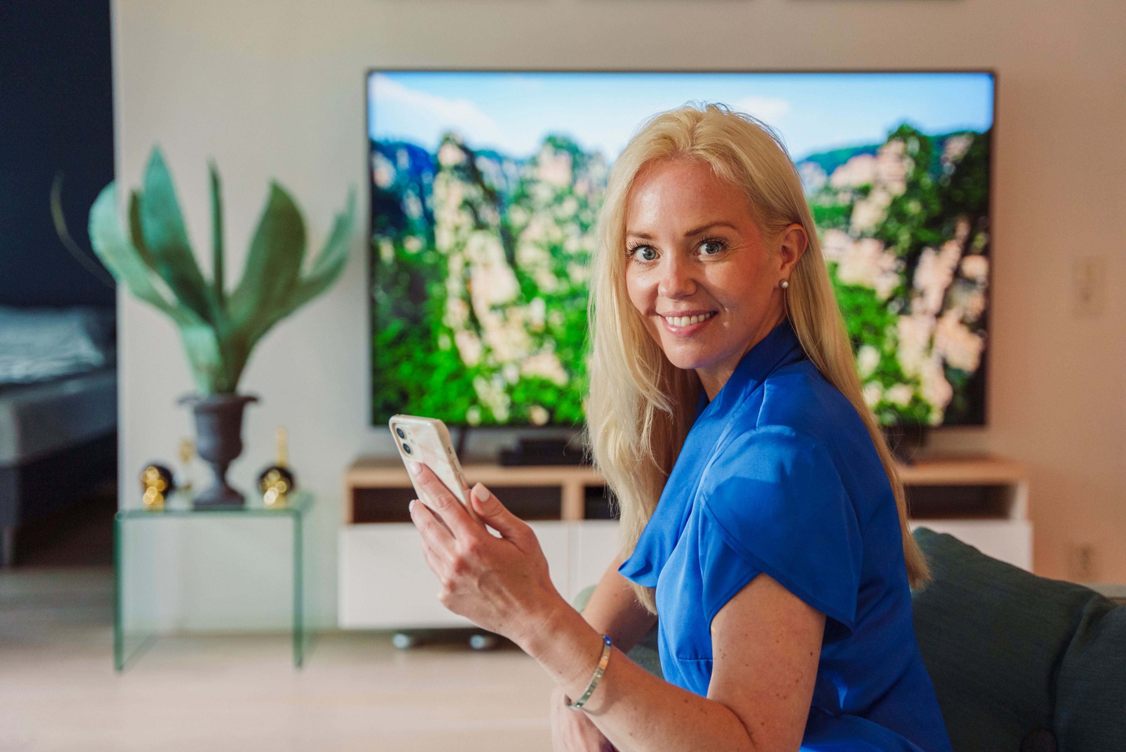 Ei dame sitter på en sofa med mobilen i hånda. Hun ser i kameraet og smiler.