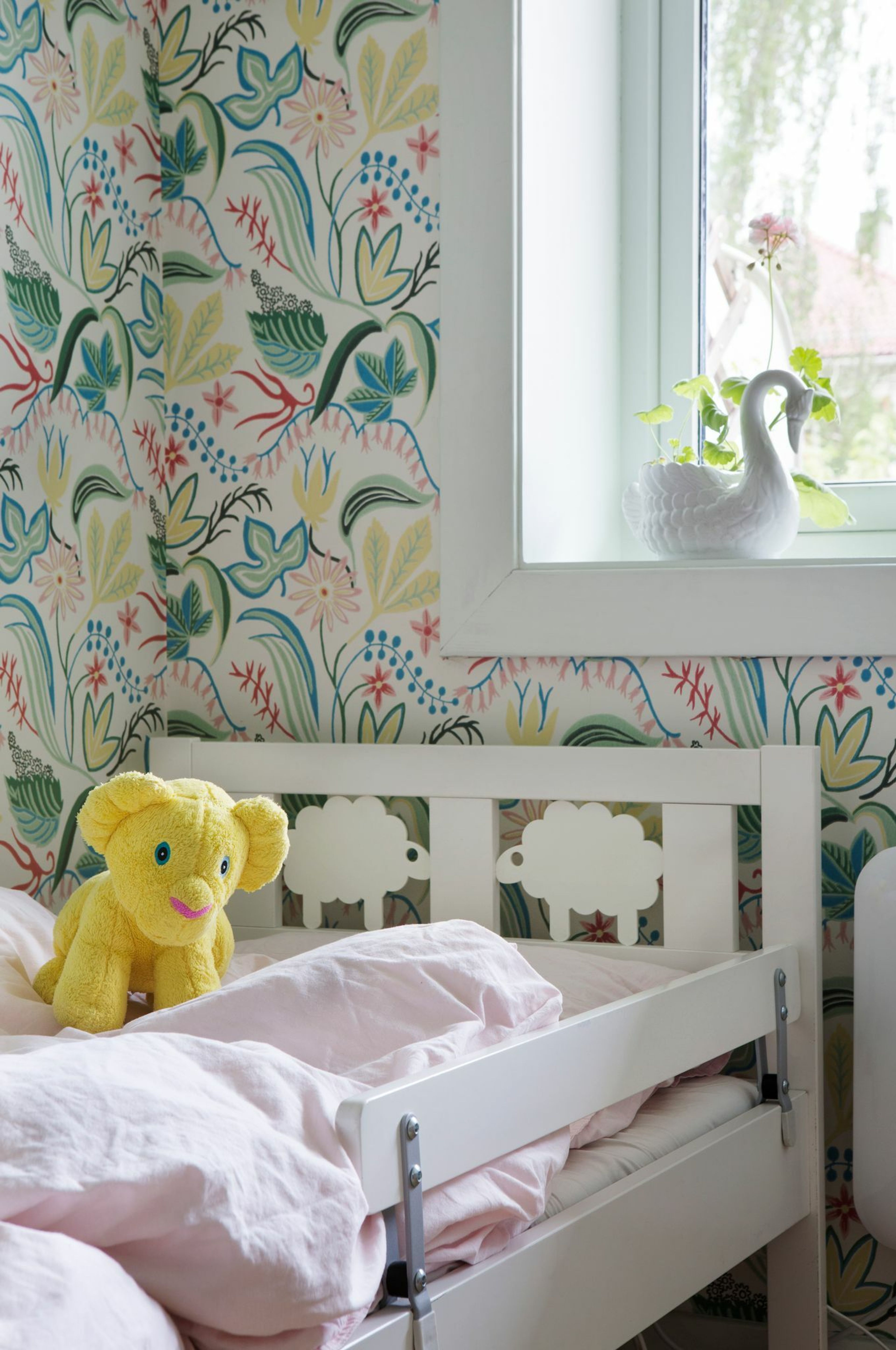 Bilde av et tapetsert barnerom med hvit barneseng med en gul løvebamse i.