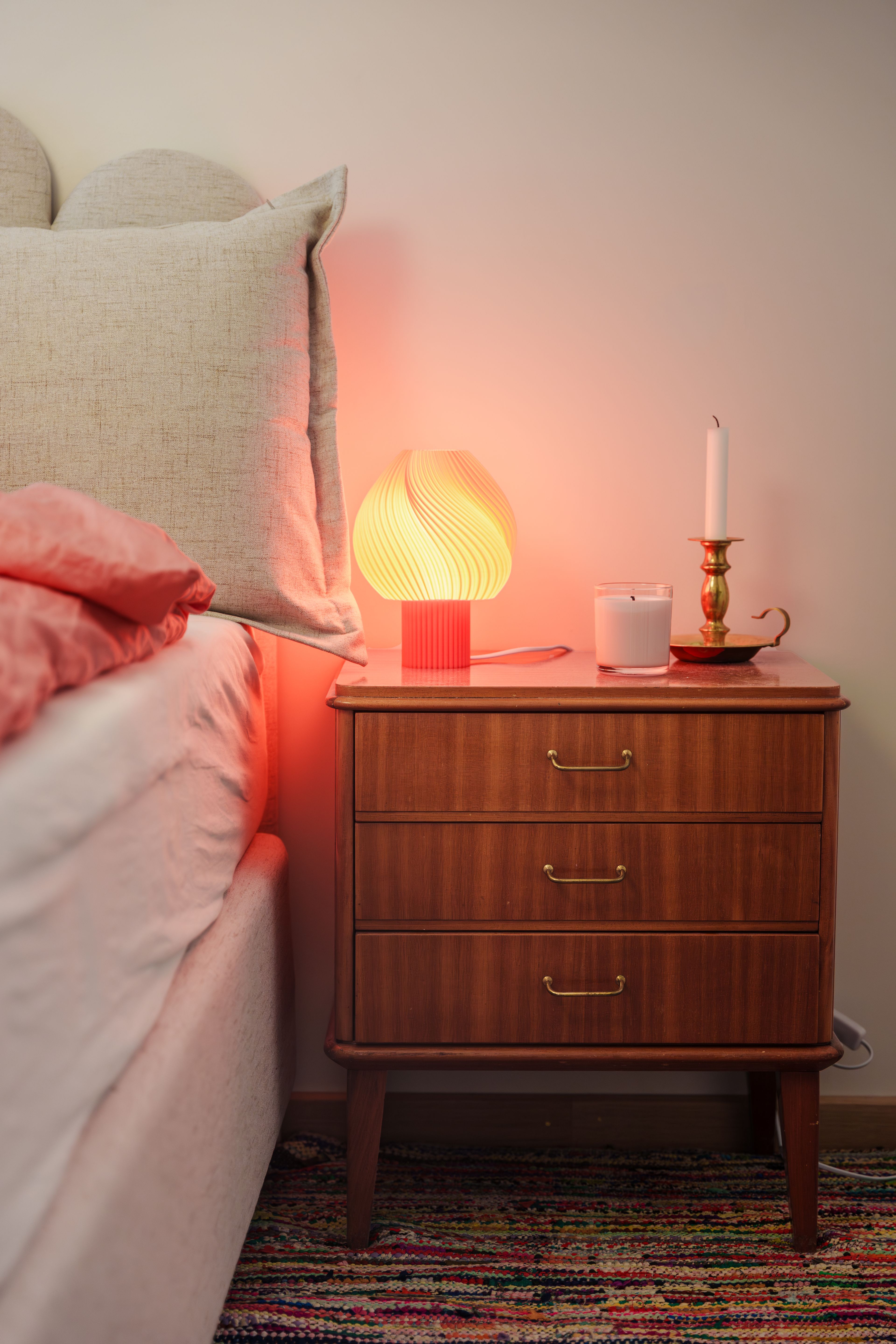 En brun kommode ved siden av senga med en rødrosa lampe oppå
