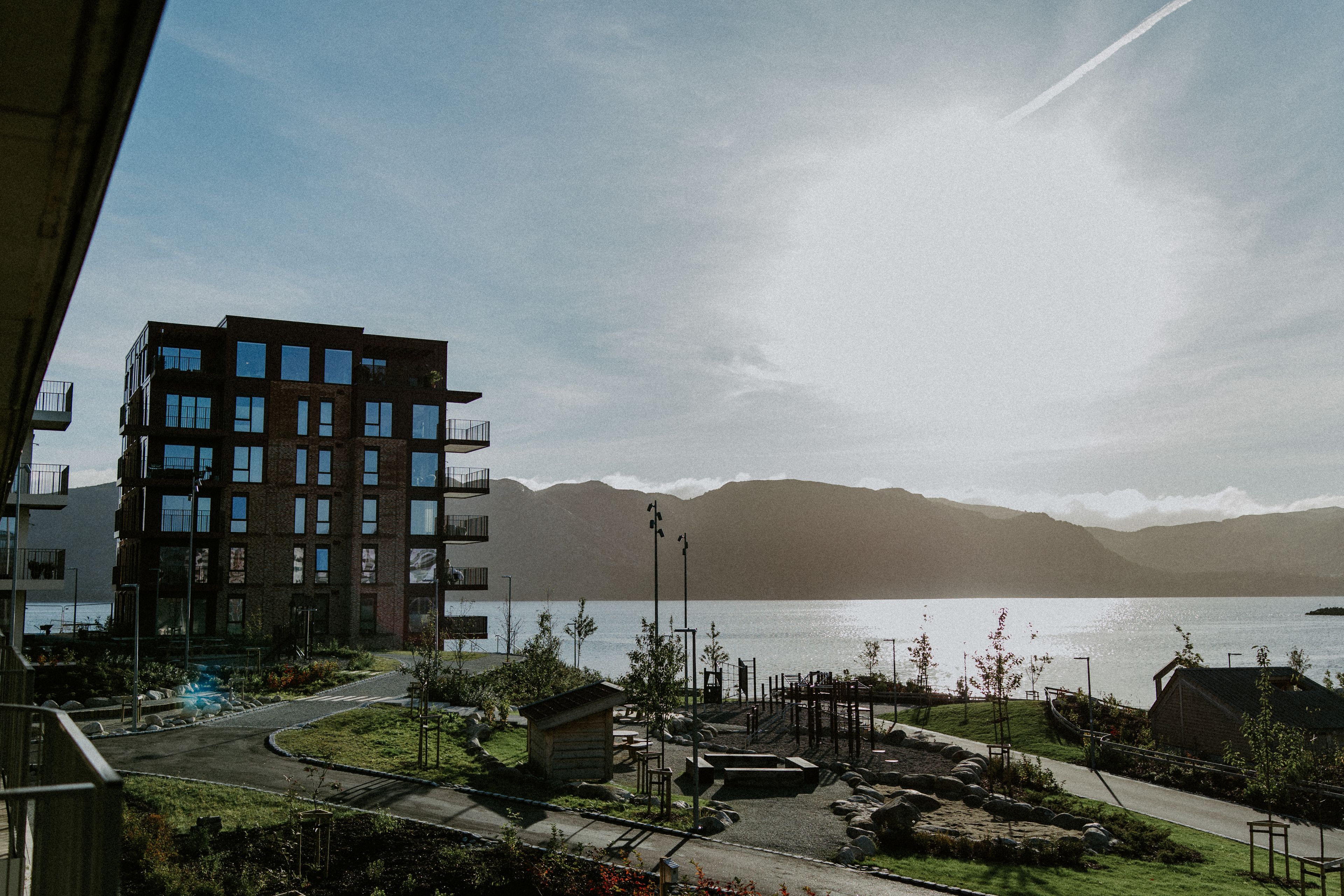 Utsikt fra en balkong på Odden mot en rød teglsteinsblokk, park med lekeplass og sjøen med fjell bak.