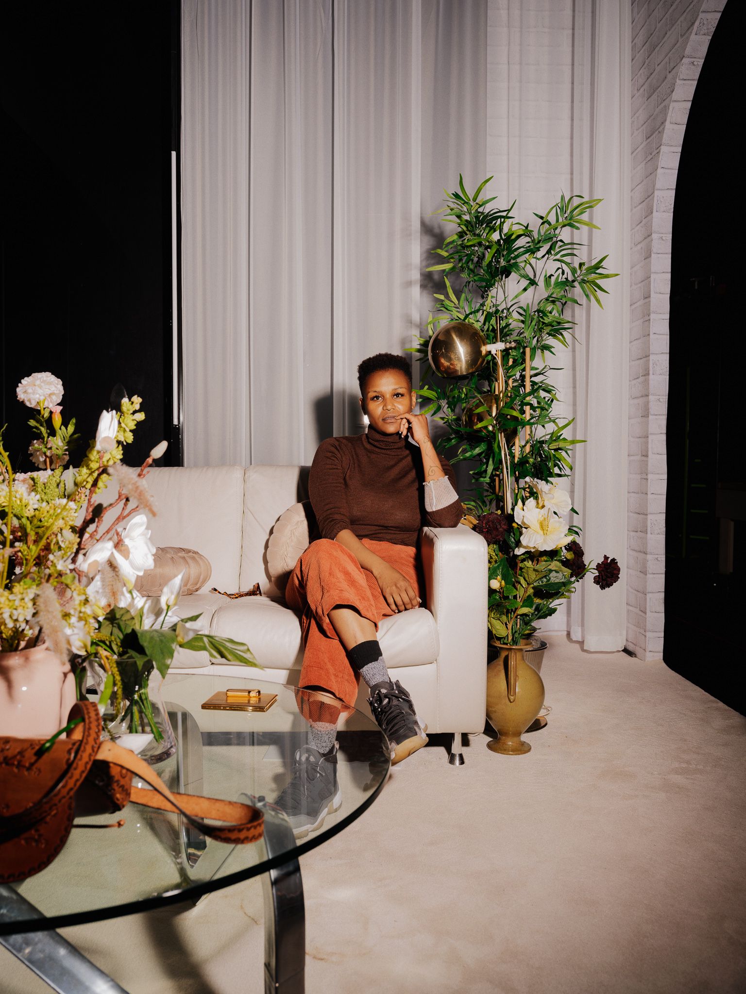 Jae Nyamburah har på seg en brun genser og oransje bukse, solbriller med rødt glass. Hun sitter i en sofa og holder hånden under haka.