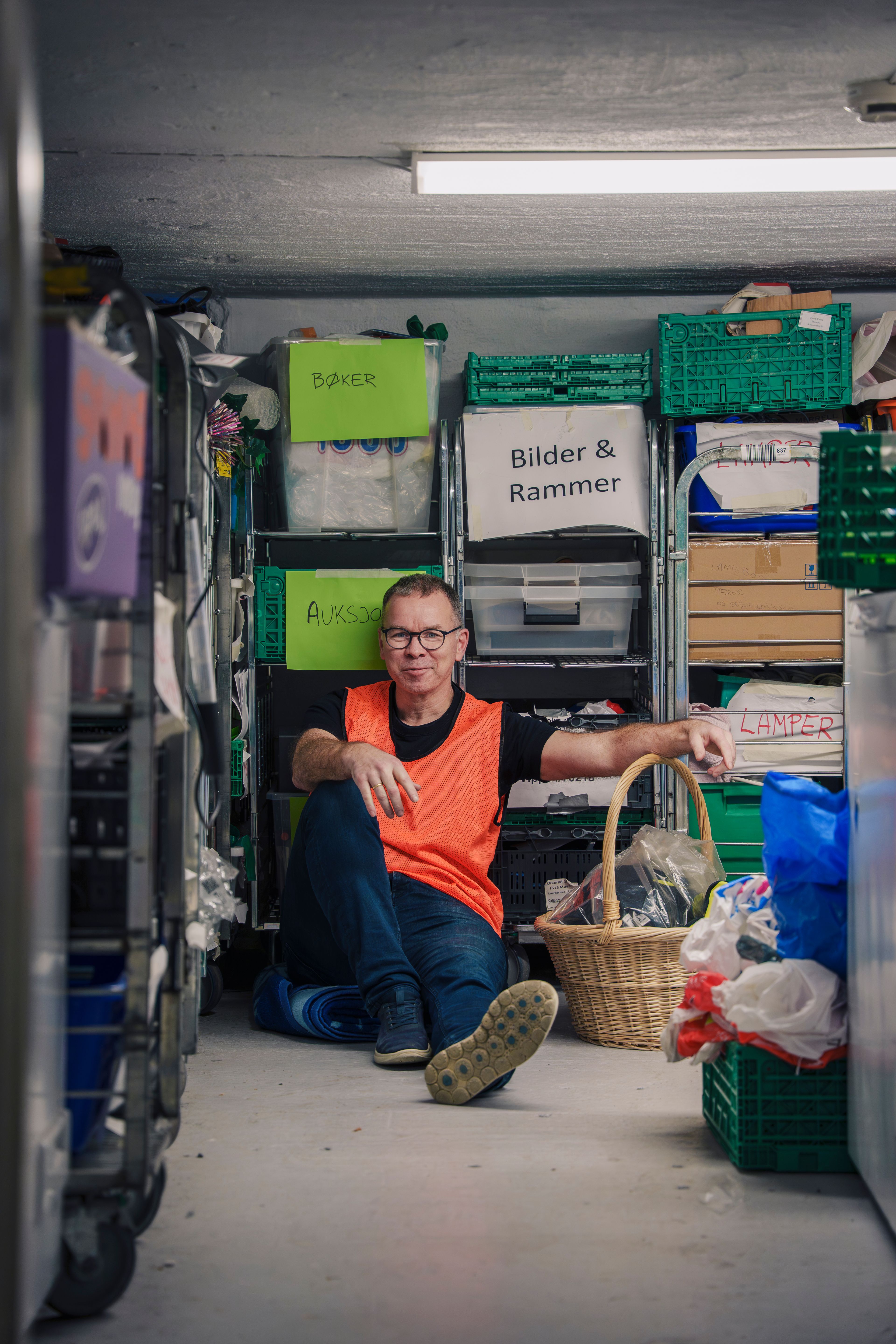 Per Christian Kittilsen sitter på gulvet i et lopperom omgitt av merkede pallehyller, kurver og pappesker.