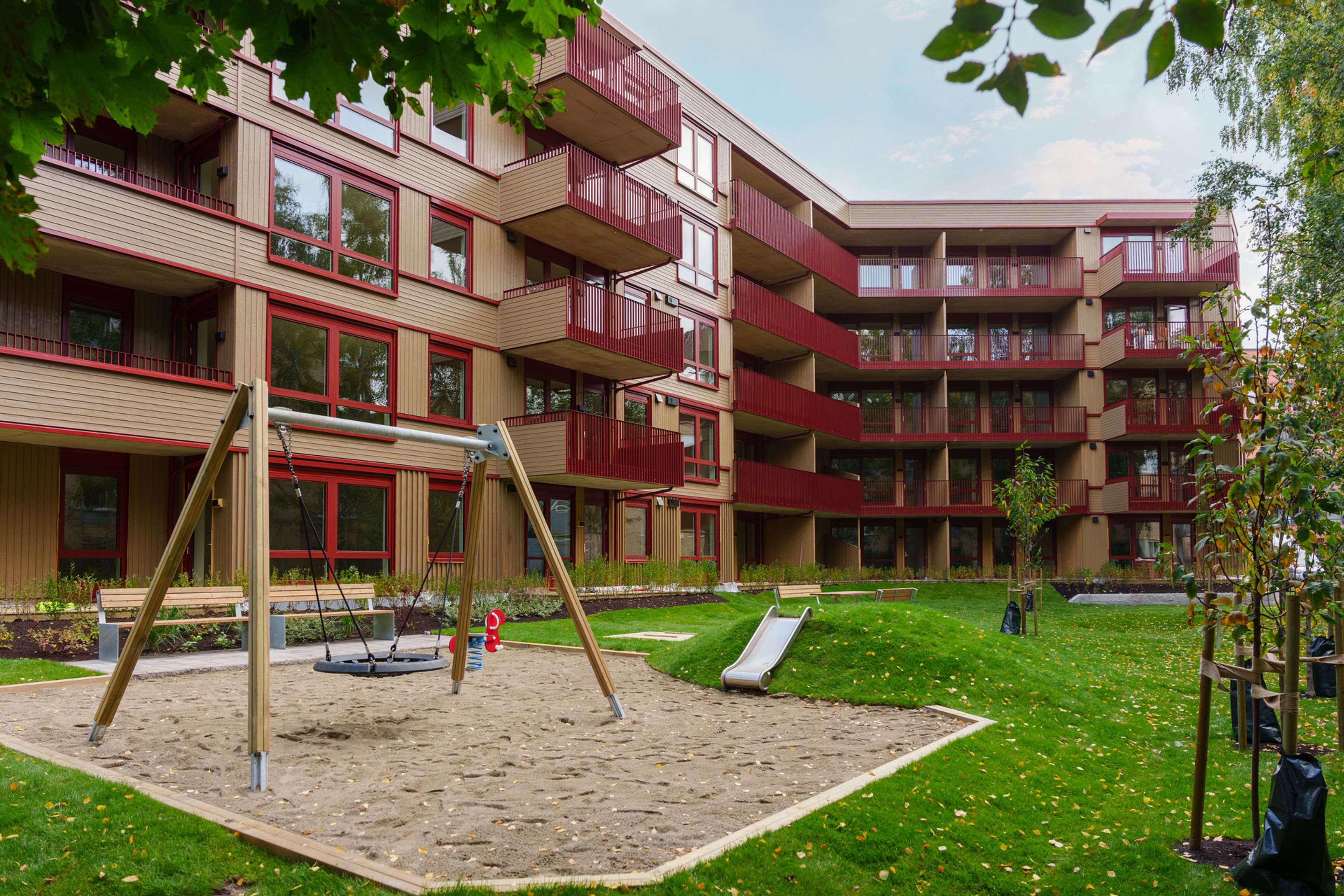 Foto av bakgården til Ensjøkroken. Det er en lekeplass av sand med en huske og en sklie. Lekeplassen er omringet av en gressplen med flere benker.
