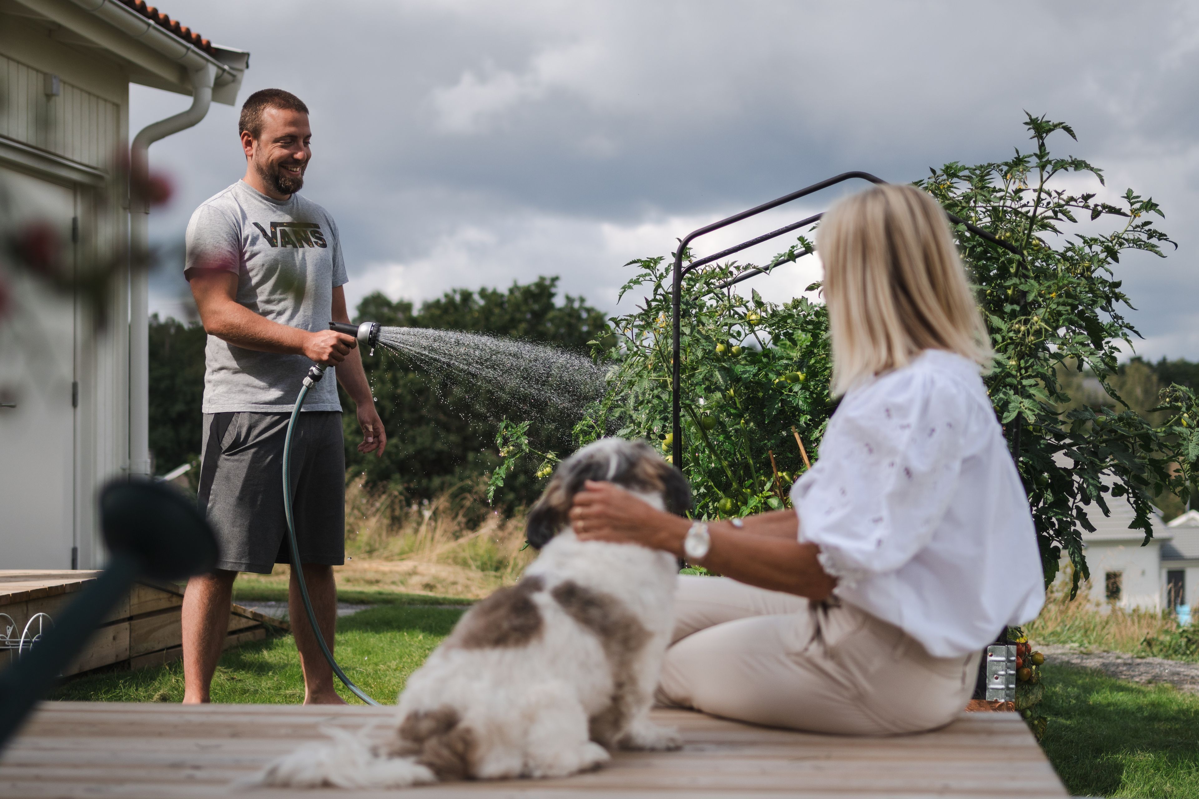 Bild på kvinna som klappar sin hund på altanen och en man som vattnar rabatten