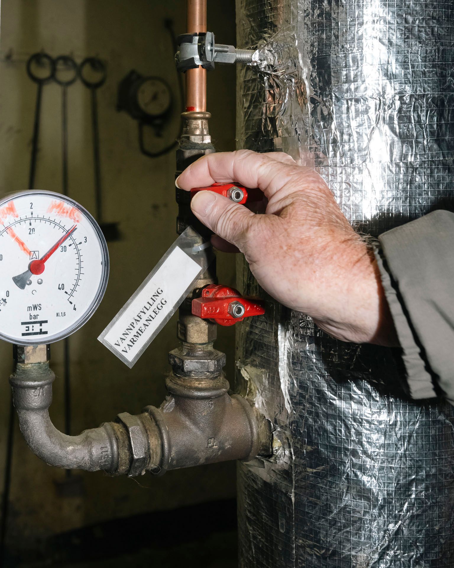 En person justerer en ventil merket ‘Vannpåfylling varmeanlegg’ ved siden av et manometer som viser trykket i rørsystemet.