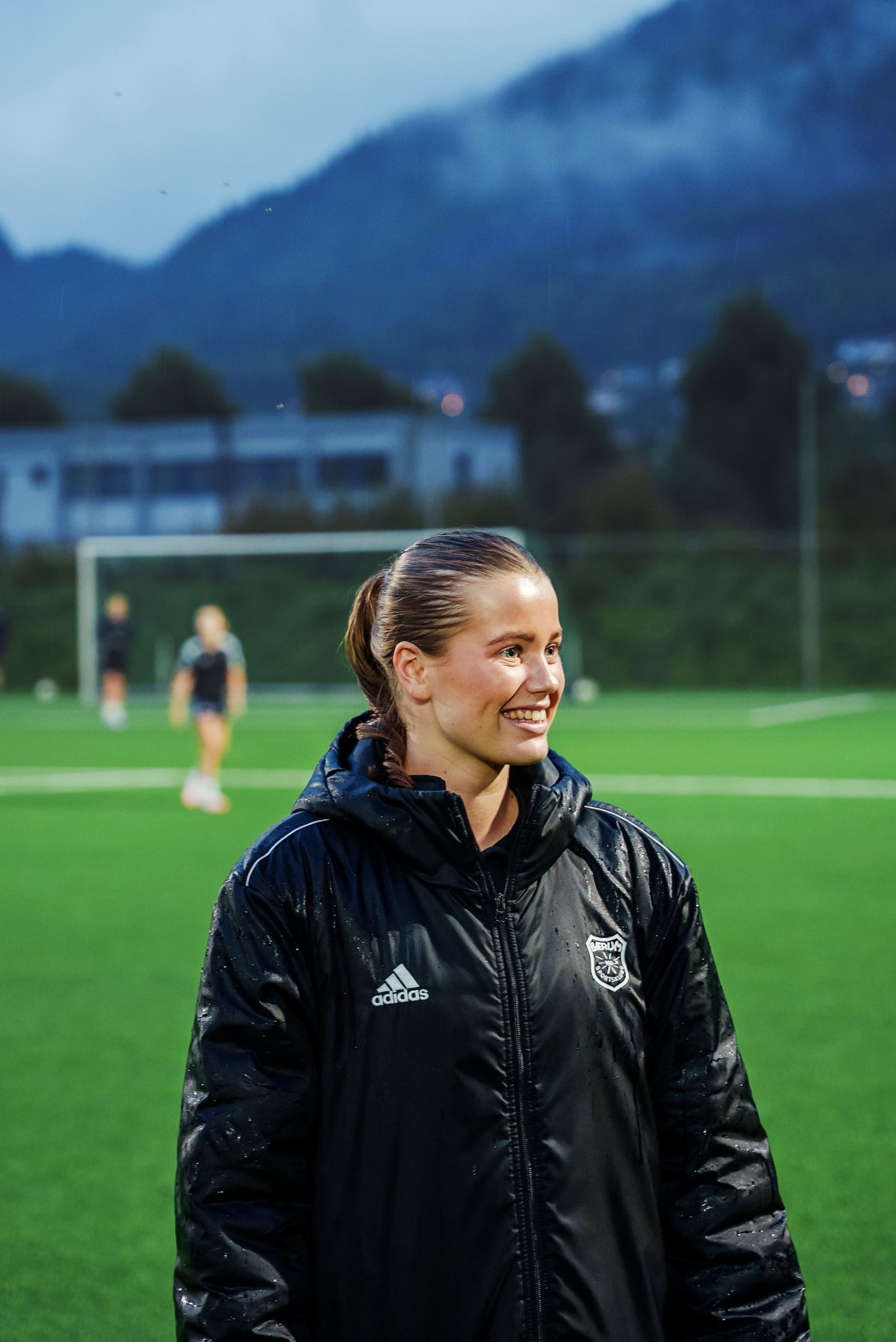 Assistenttrener Hedda smiler og ser til høyre for kamera med fotballbanen i bakgrunnen.