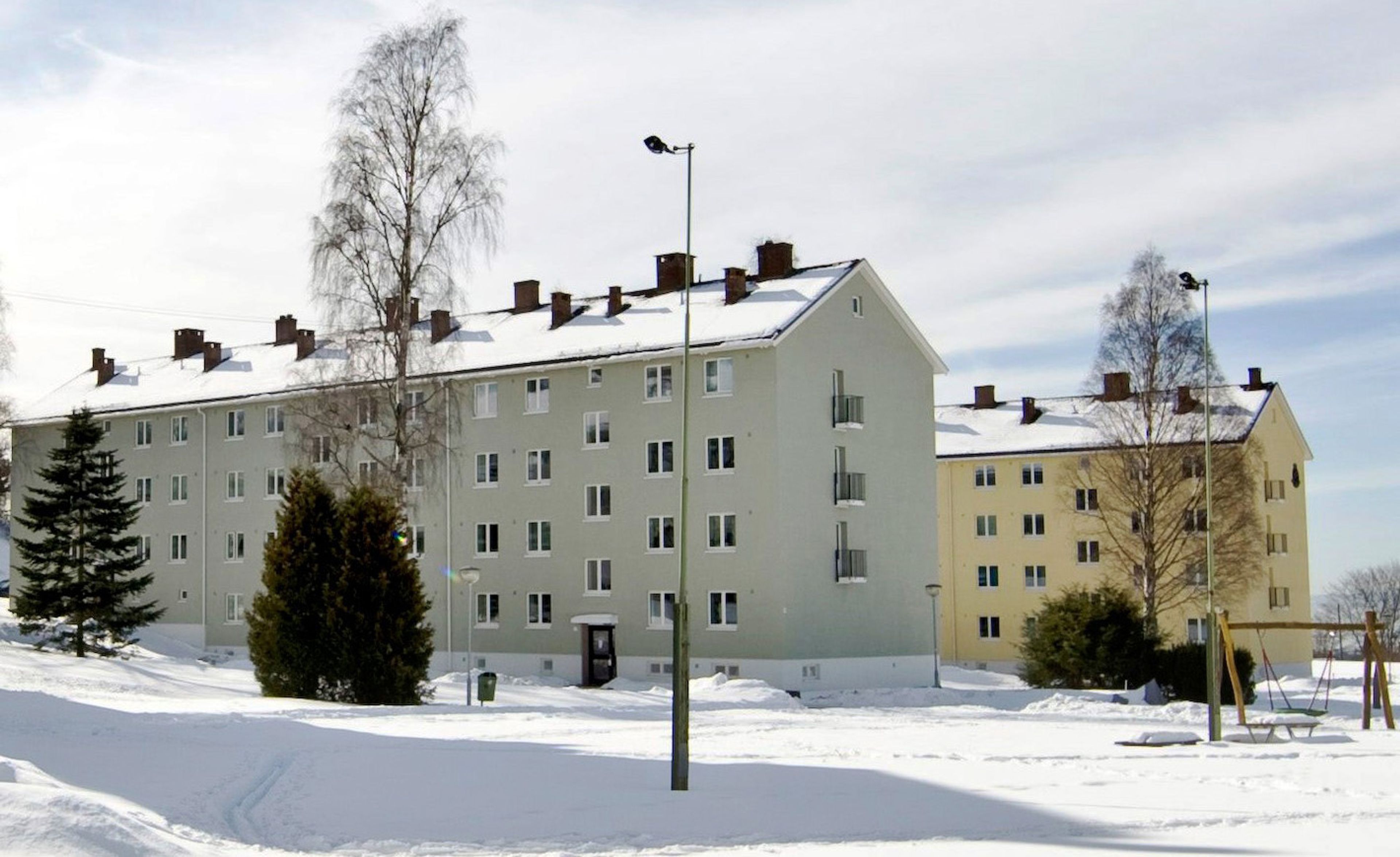 En grå boligblokk, med en gul blokk i bakgrunnen, på vintertid.