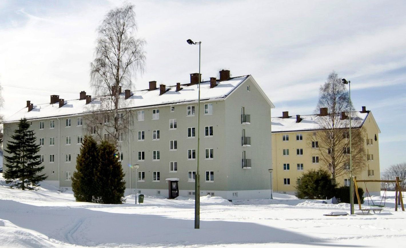 En grå boligblokk, med en gul blokk i bakgrunnen, på vintertid.