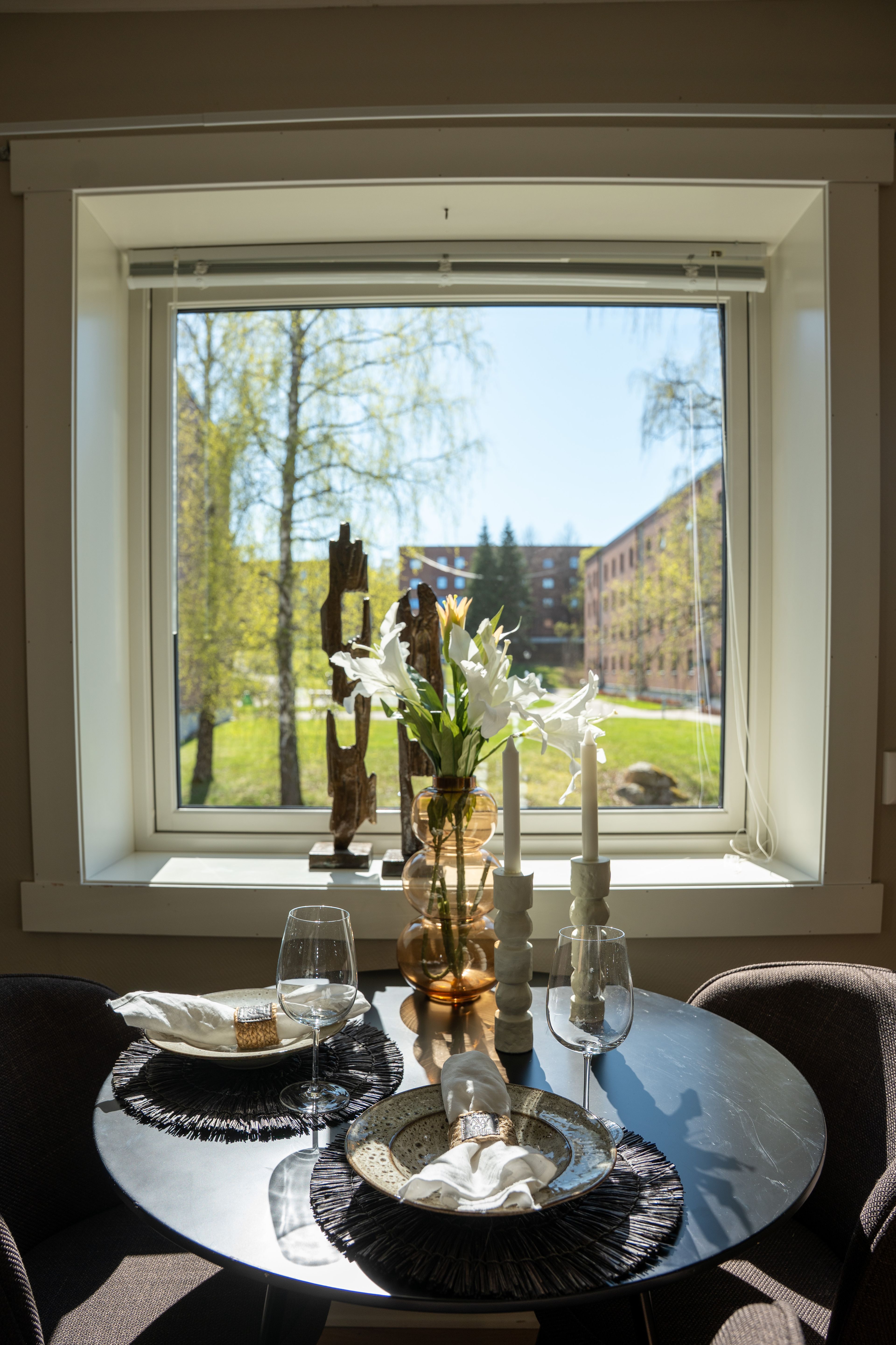 undt mørkt bord dekket til to, med blomster og stearinlys. Vindu med utsikt til trær og rød murbygning i bakgrunnen.