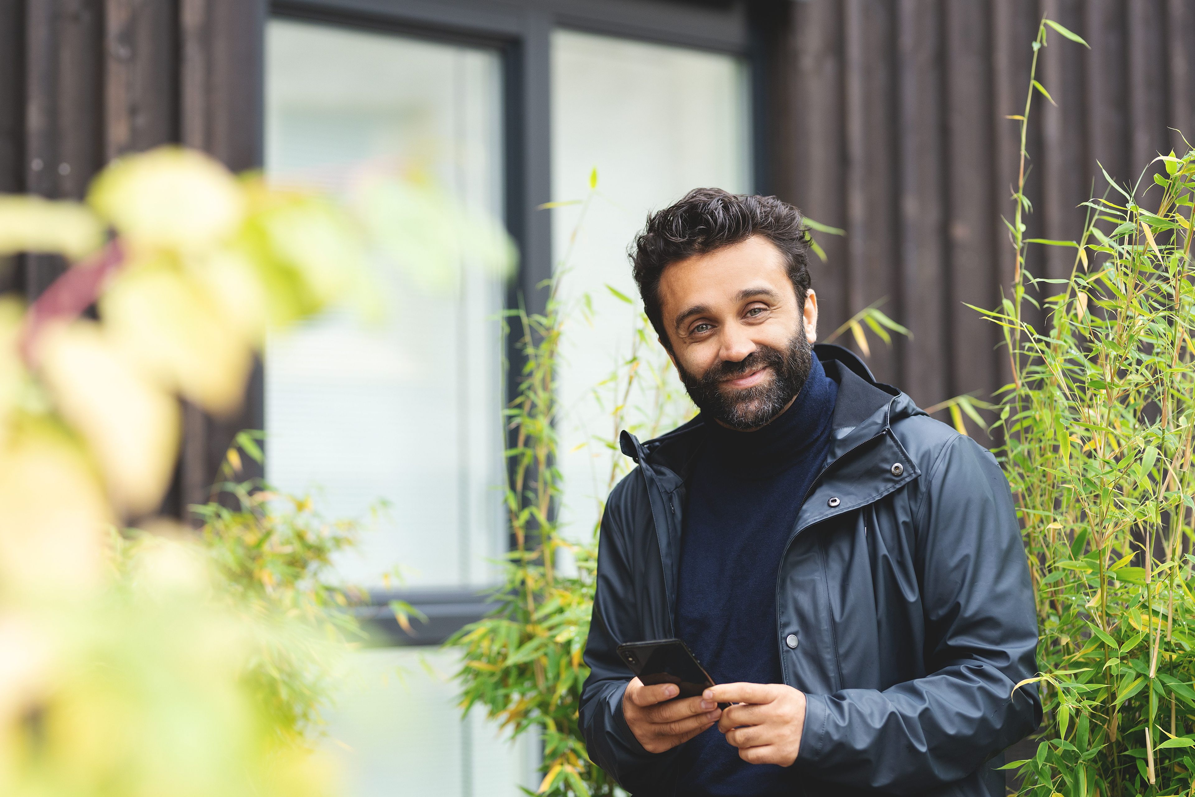 En mann ser mot kamera og smiler. Han står utenfor et hus. Det er grønne busker rundt og han holder en mobiltelefon i hånda.