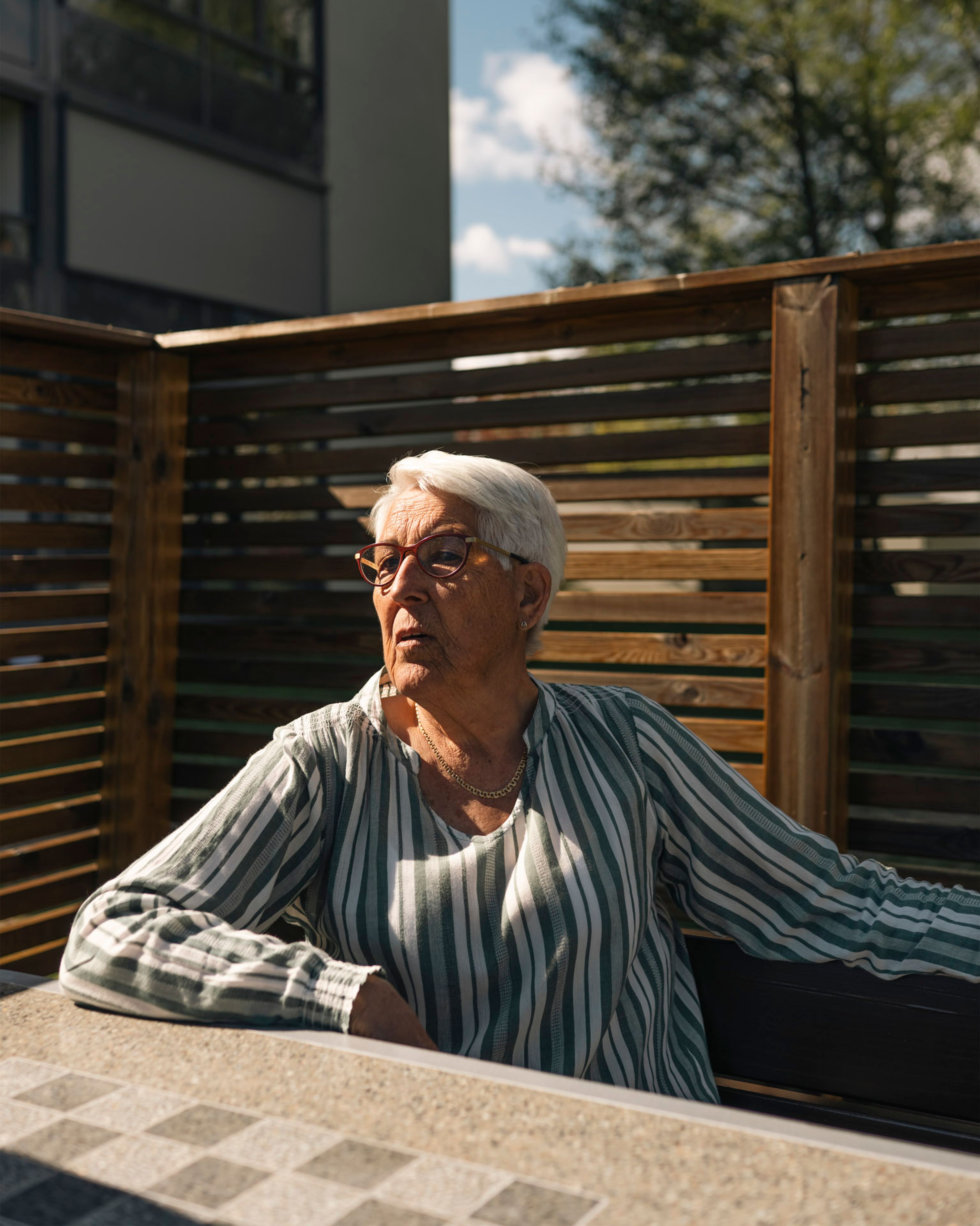 Eldre kvinne sitter ute i solen ved et bord foran en levegg i tre i et boligområde.