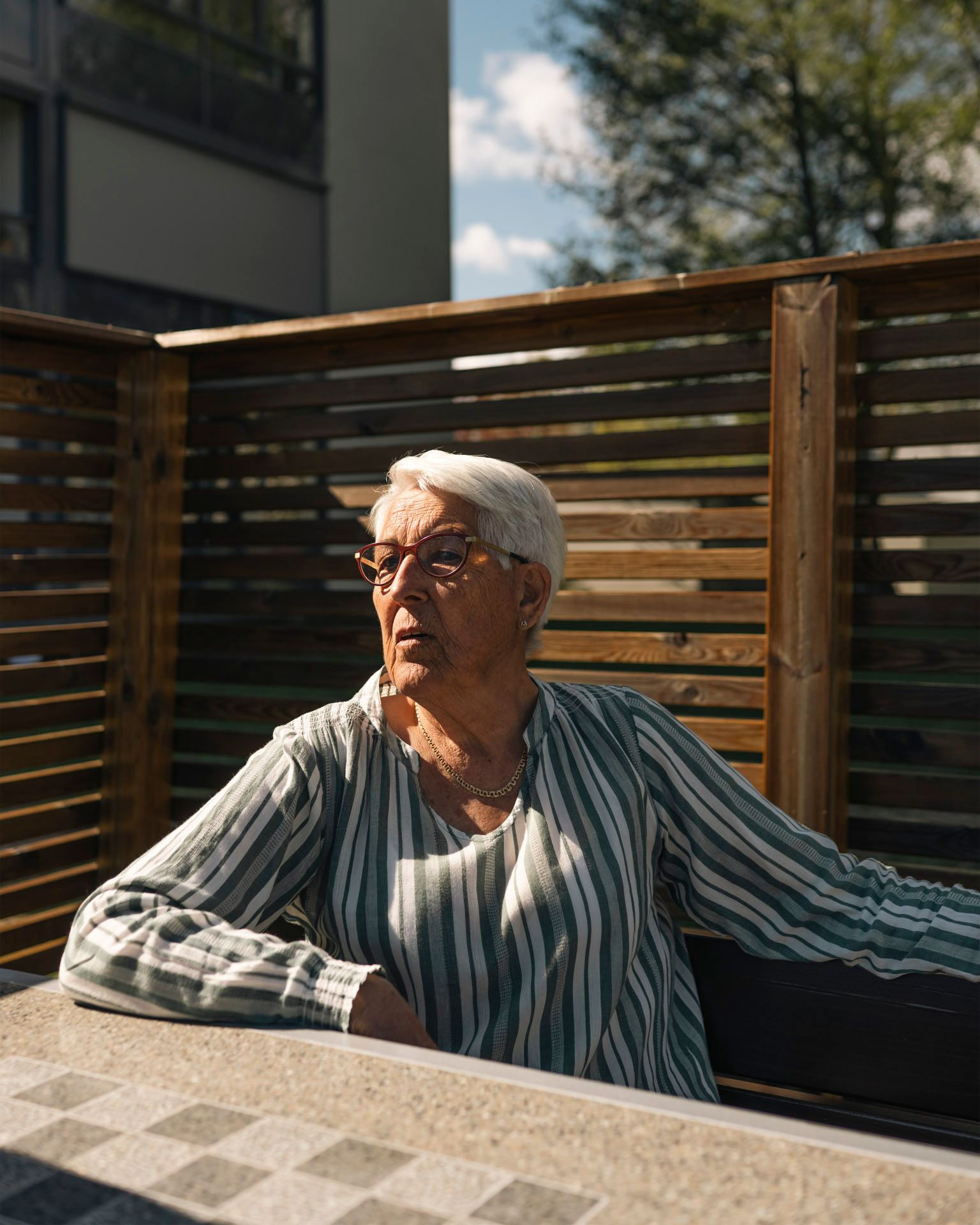 Eldre kvinne sitter ute i solen ved et bord foran en levegg i tre i et boligområde.