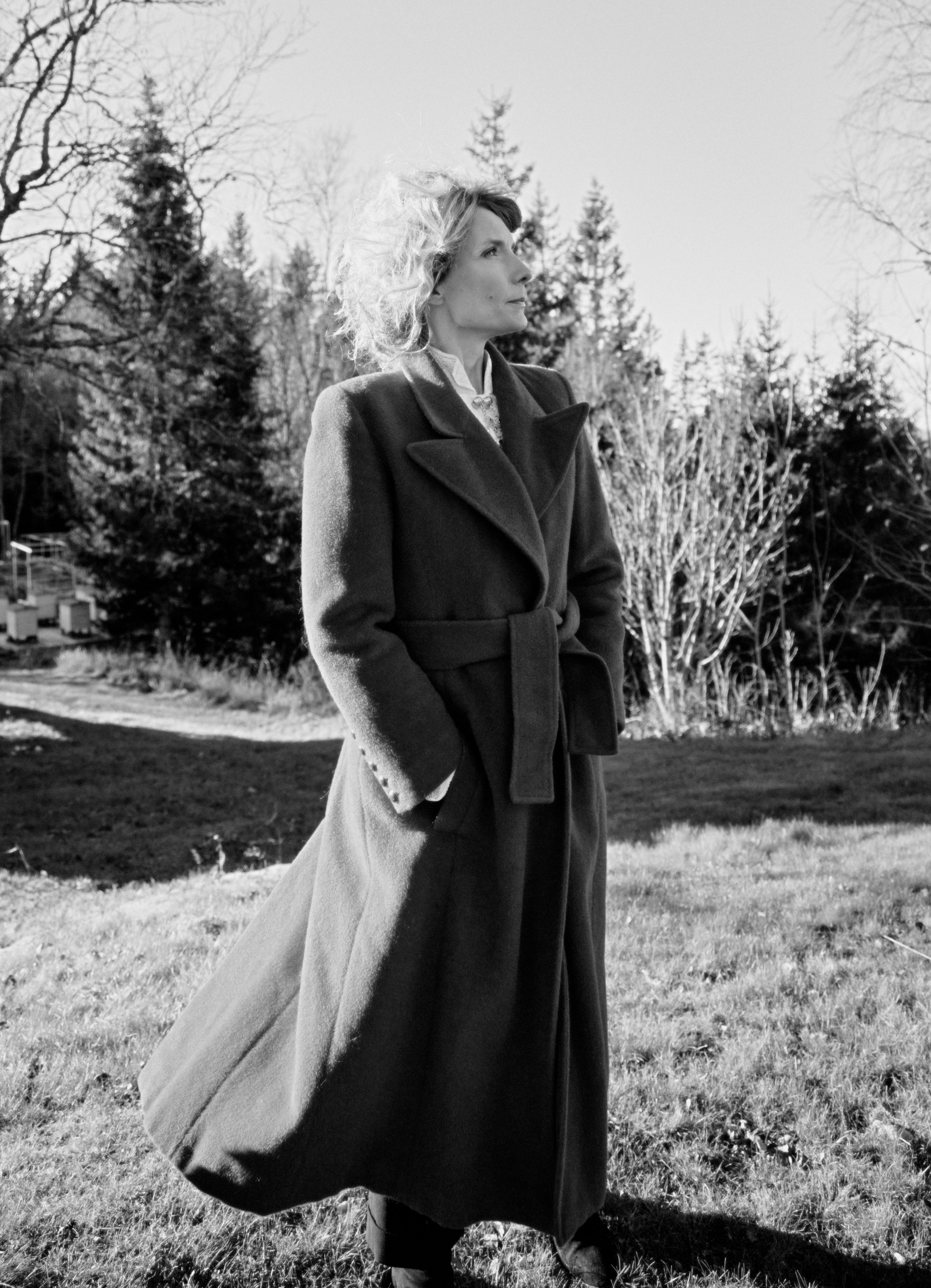 Et svart/hvitt foto av Herborg Kråkvik i helfigur. Hun står på en gresslette i frakk, med hendene i lomma og ser mot venstre.