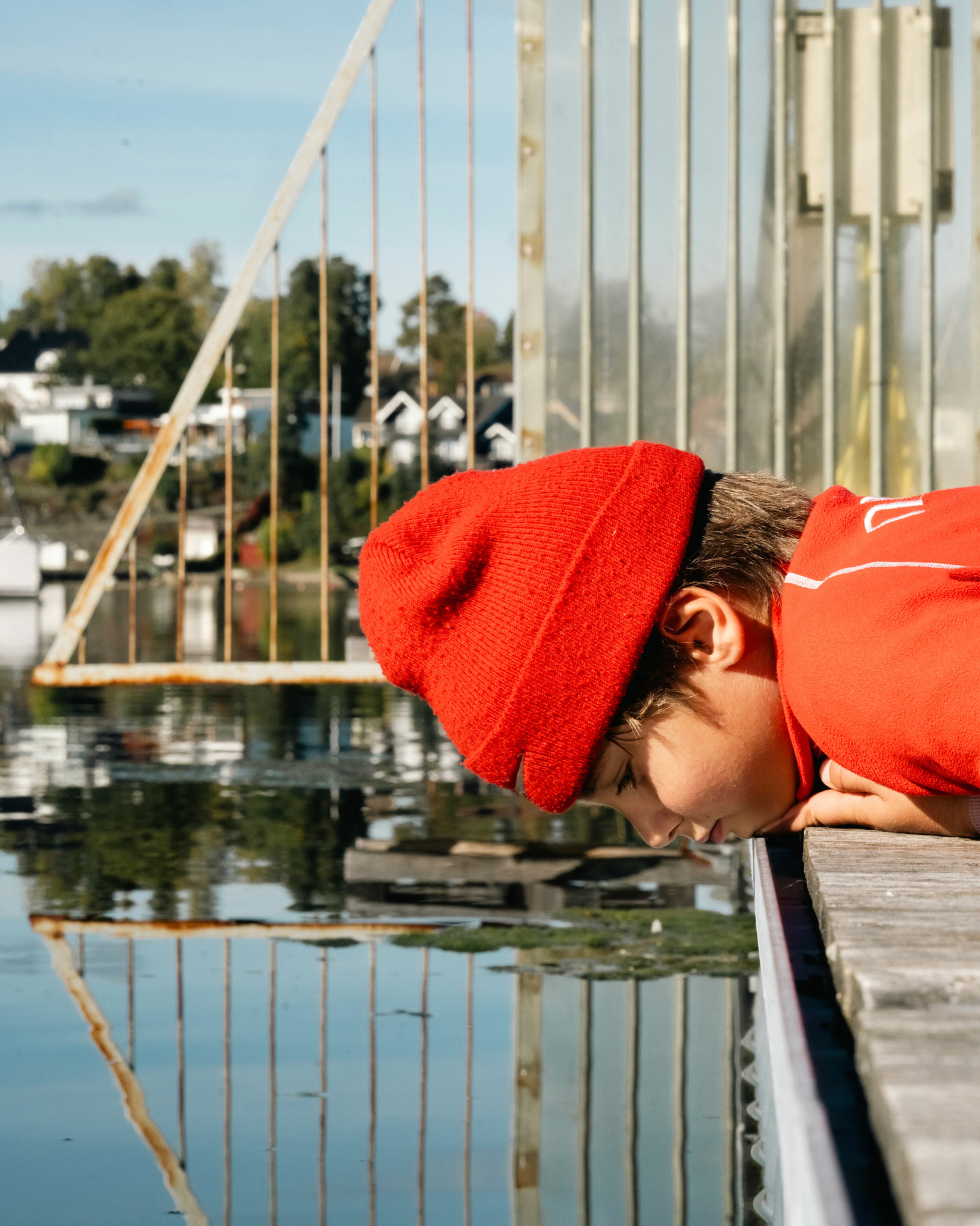 En gutt med en rød lue ligger på en brygge og titter ned i vannet.