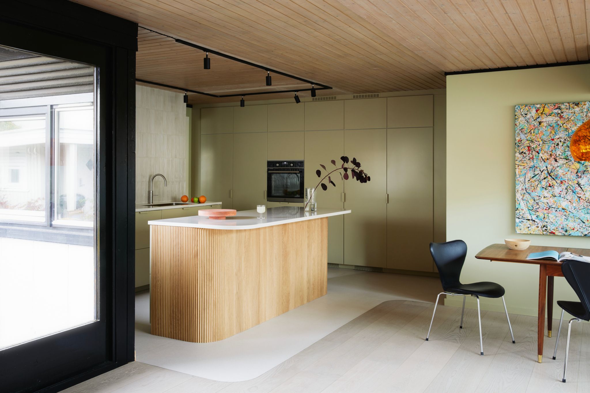 Et moderne kjøkken med en lys treøy og olivengrønne skap i bakgrunnen. Ved siden av kjøkkenet er det et spisebord med svarte stoler og et fargerikt kunstverk på veggen.