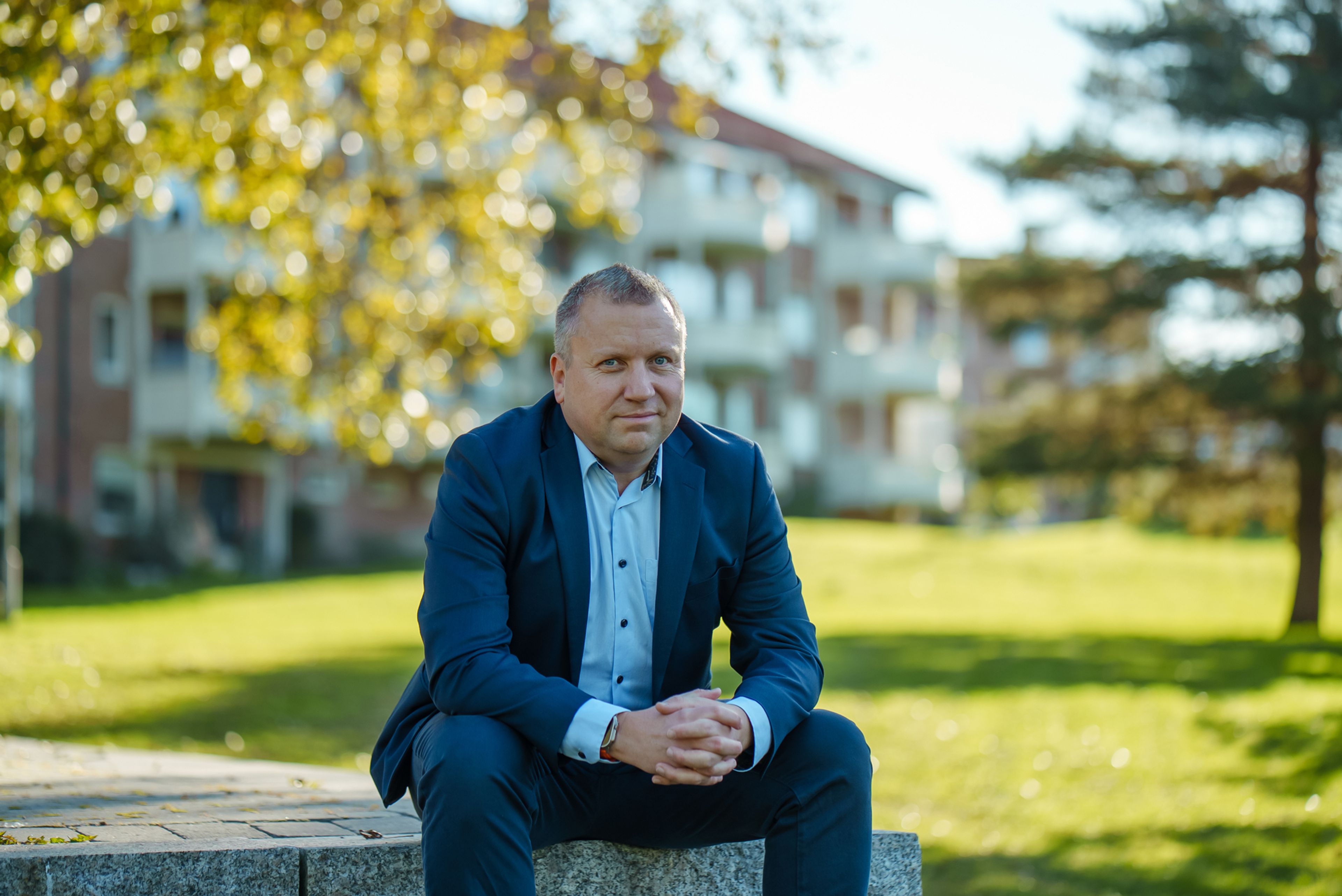 Øistein Gamst Sandlie sitter på steiner og folder hendene i fanget. Han smiler. Han sitter utenfor OBOS-blokk. Det er grønt g frodig bak ham, og solen skinner. 