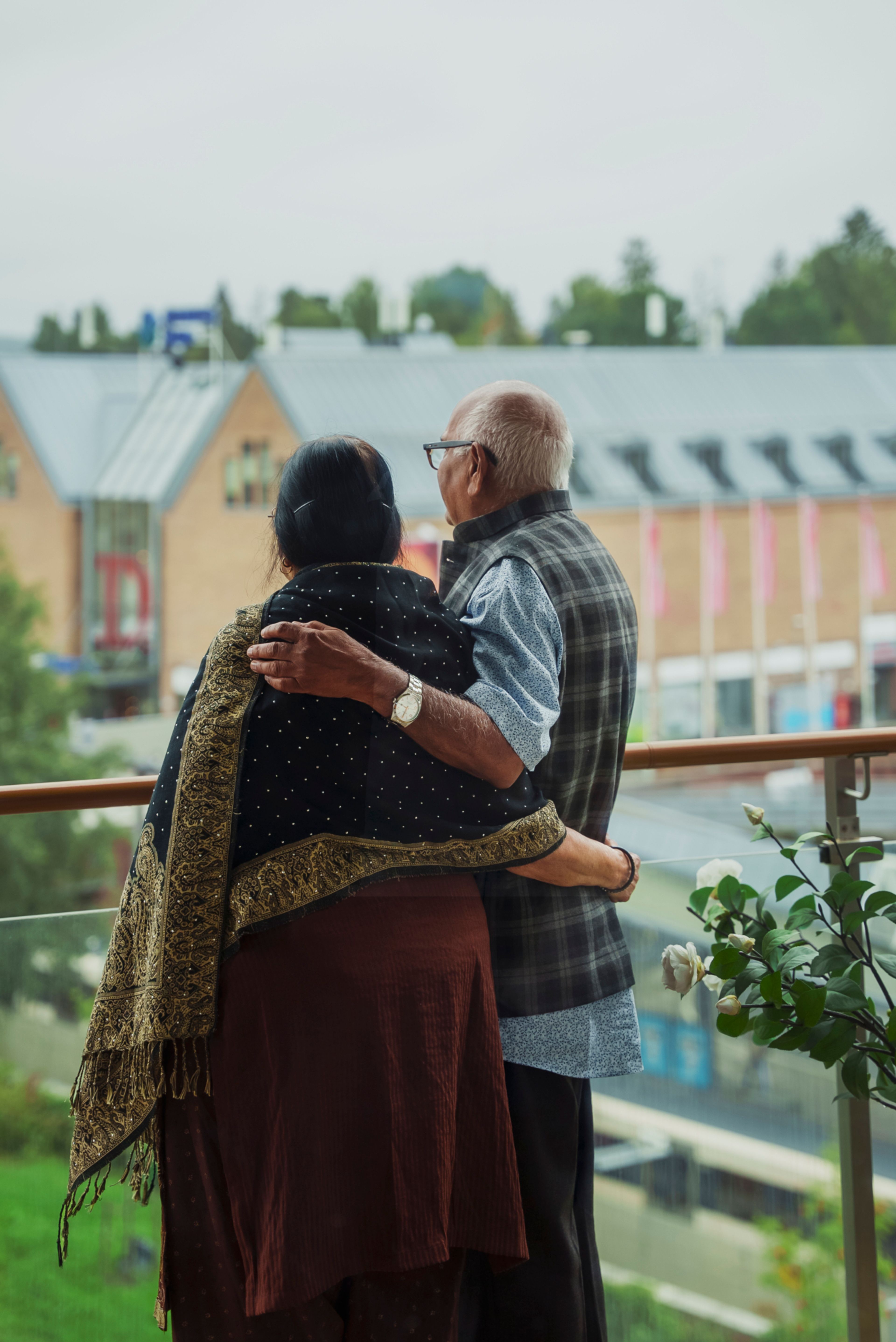 Et eldre ektepar holder armene over skulderne til hverandre, mens de står på balkongen og ser utover bakgården sin.
