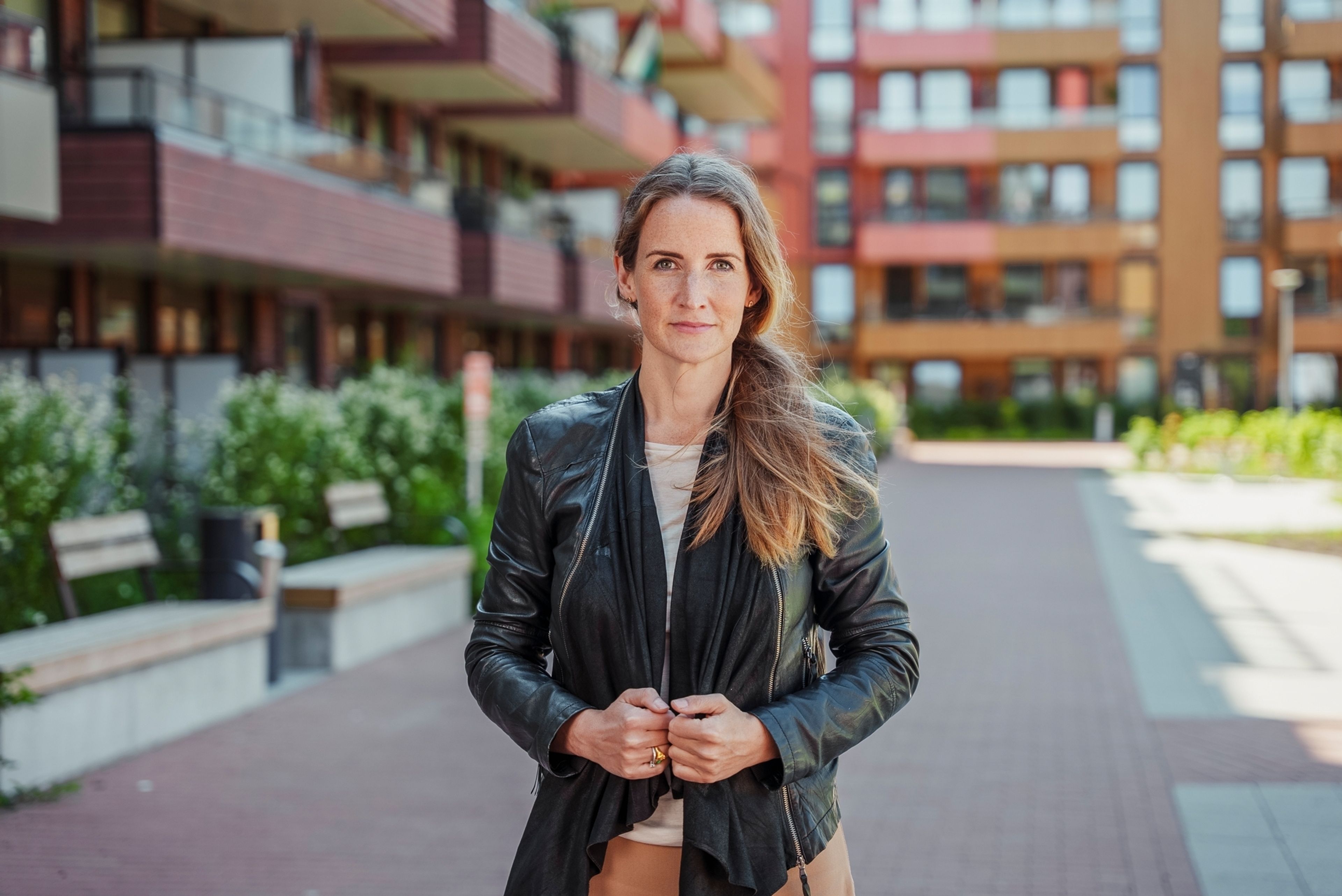 Ingjerd står og smiler forsiktig mot kamera i en svart skinnjakke, omgitt av moderne bygg i sola.