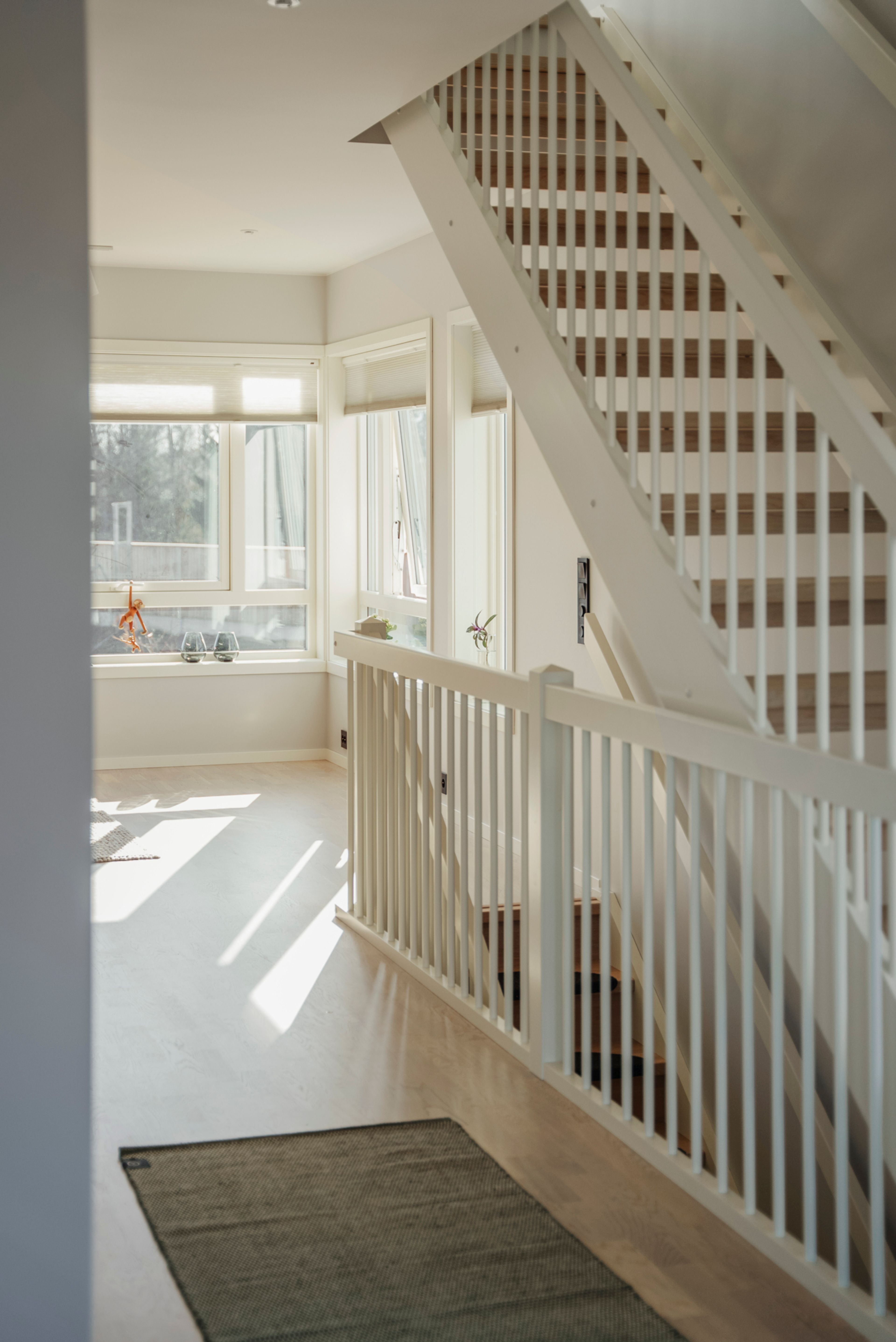 En lys stue med trappeoppgang i midten mot et par vinduer som slipper inn sol