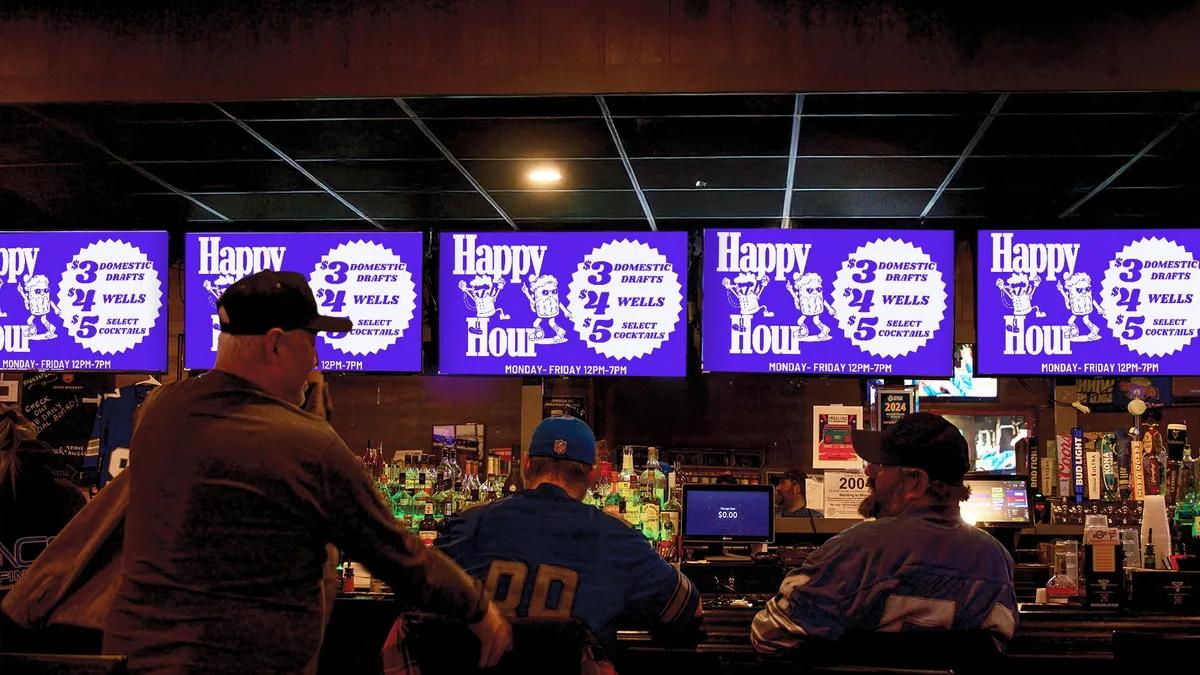 Patrons in team jerseys at a bar, with the overhead TVs running the venue's own Happy Hour drink specials
