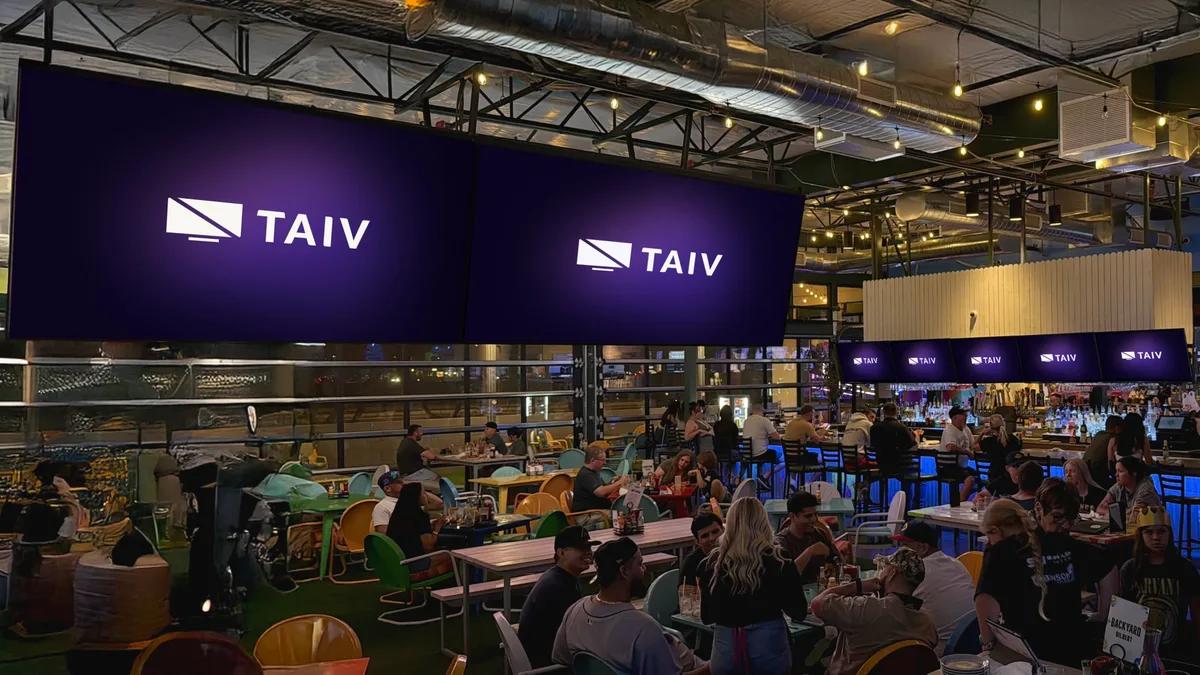A busy open-air sports bar and food hall with a wall of Taiv screens above a packed crowd