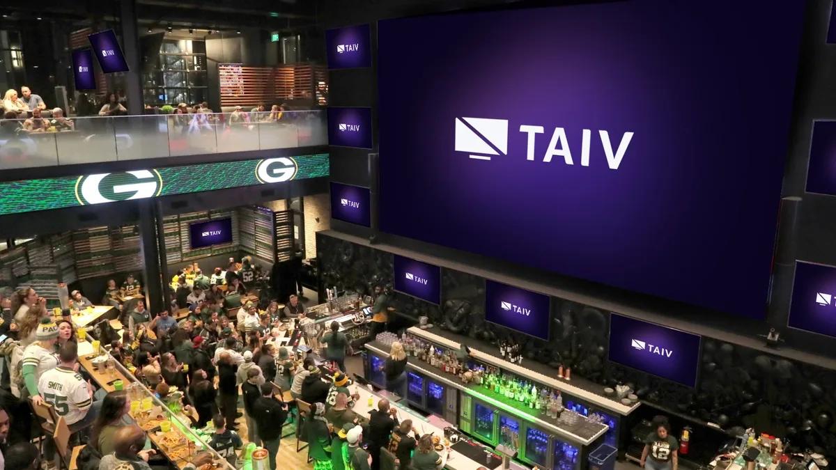 A packed multi-level sports bar on game day, Packers fans watching a huge Taiv screen above the bar