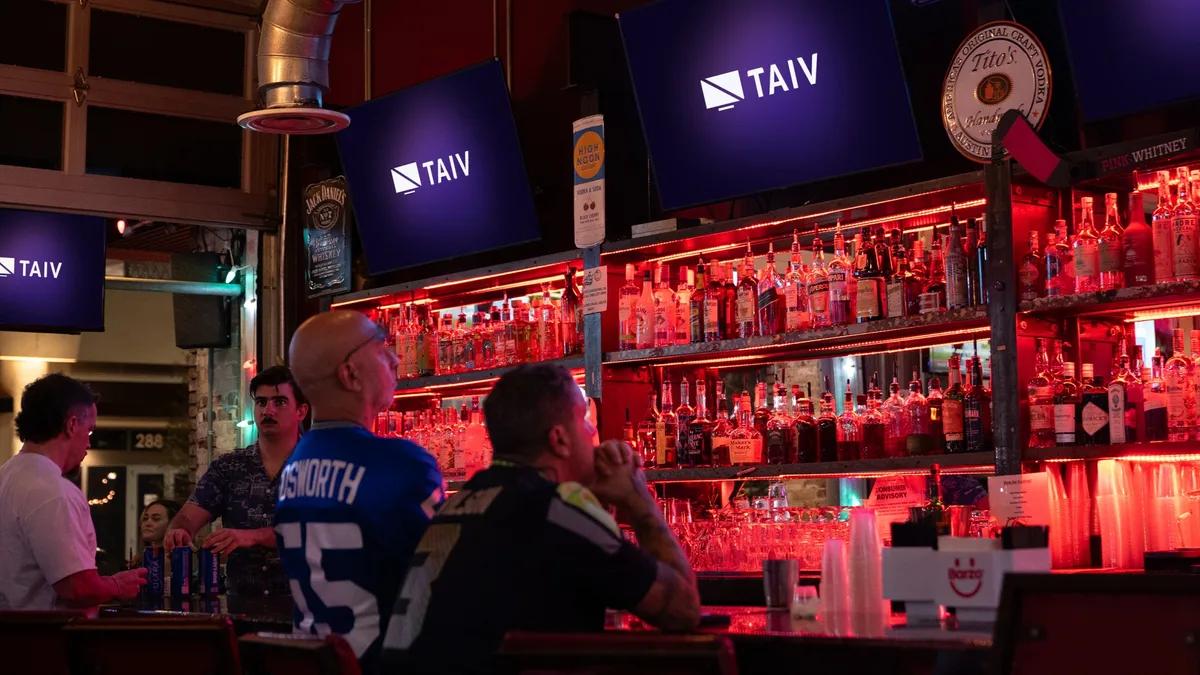 Fans in team jerseys watching Taiv screens at a red-lit bar with a full liquor back-bar